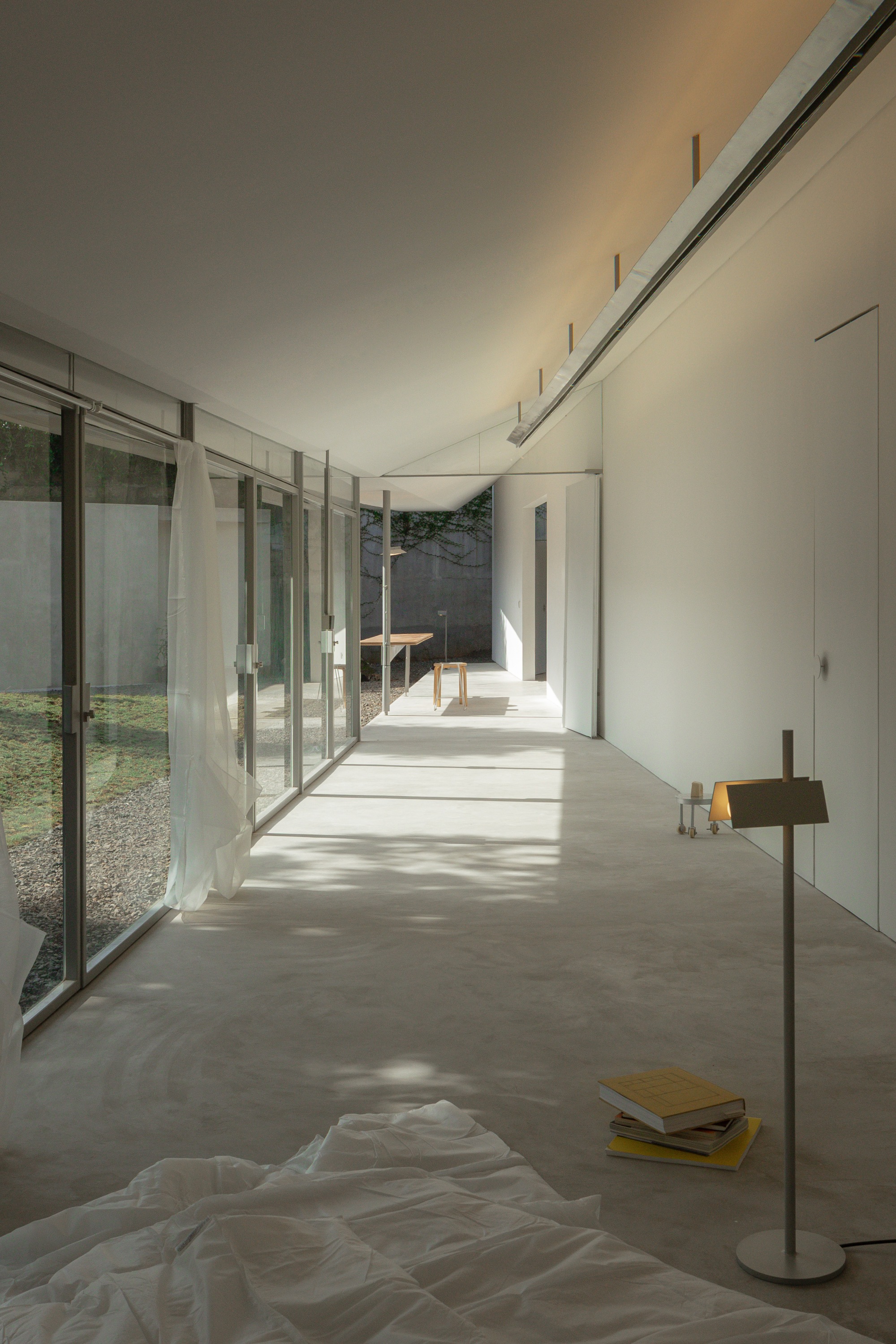 Linear interior space with glass wall, concrete floor, and draped fabric beside a reading lamp