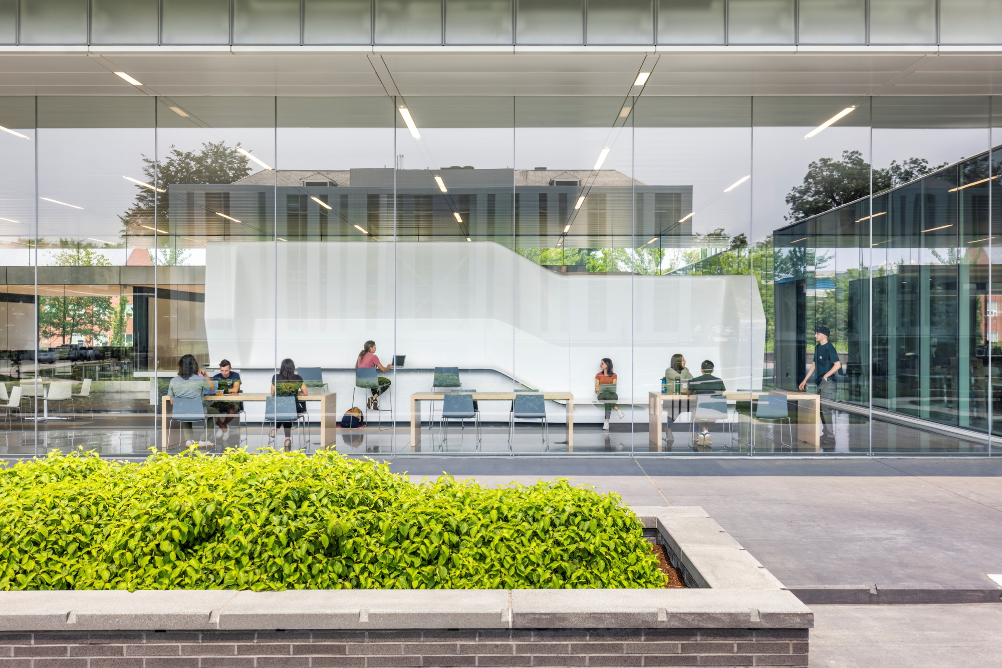 Ground-level view through floor-to-ceiling glass showing a study area with planted hedge bed in foreground
