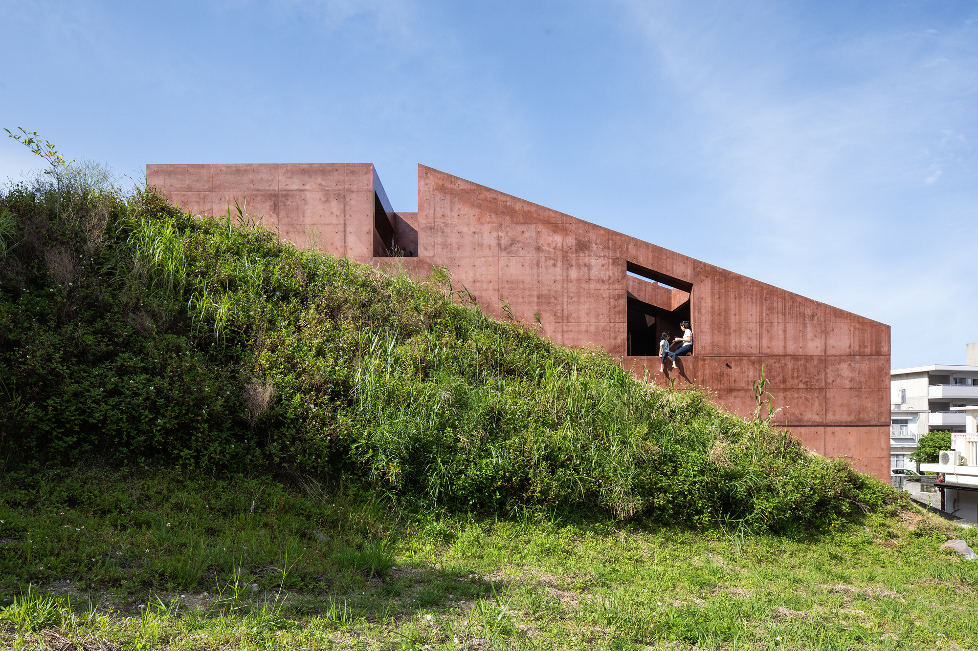 Rammed earth volumes embedded in a grassy hillside with a figure visible in an upper opening
