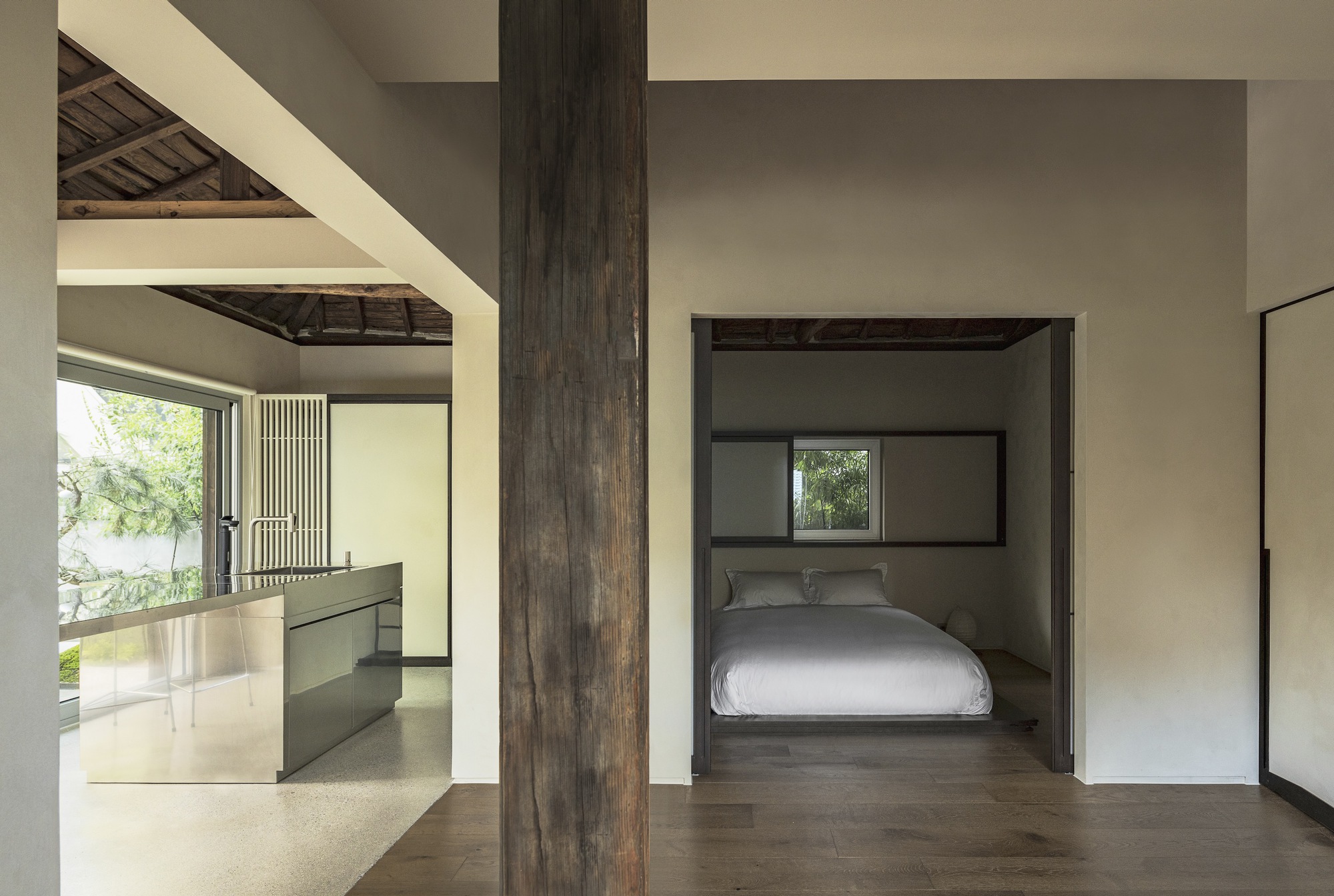 View through a dark timber column toward a bedroom alcove and kitchen with exposed roof trusses