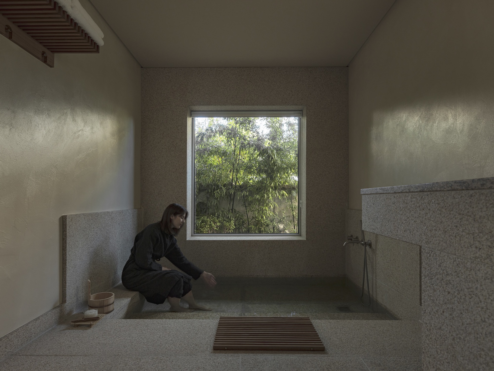 Stone-lined bathing room with a person kneeling beside the tub and window overlooking greenery