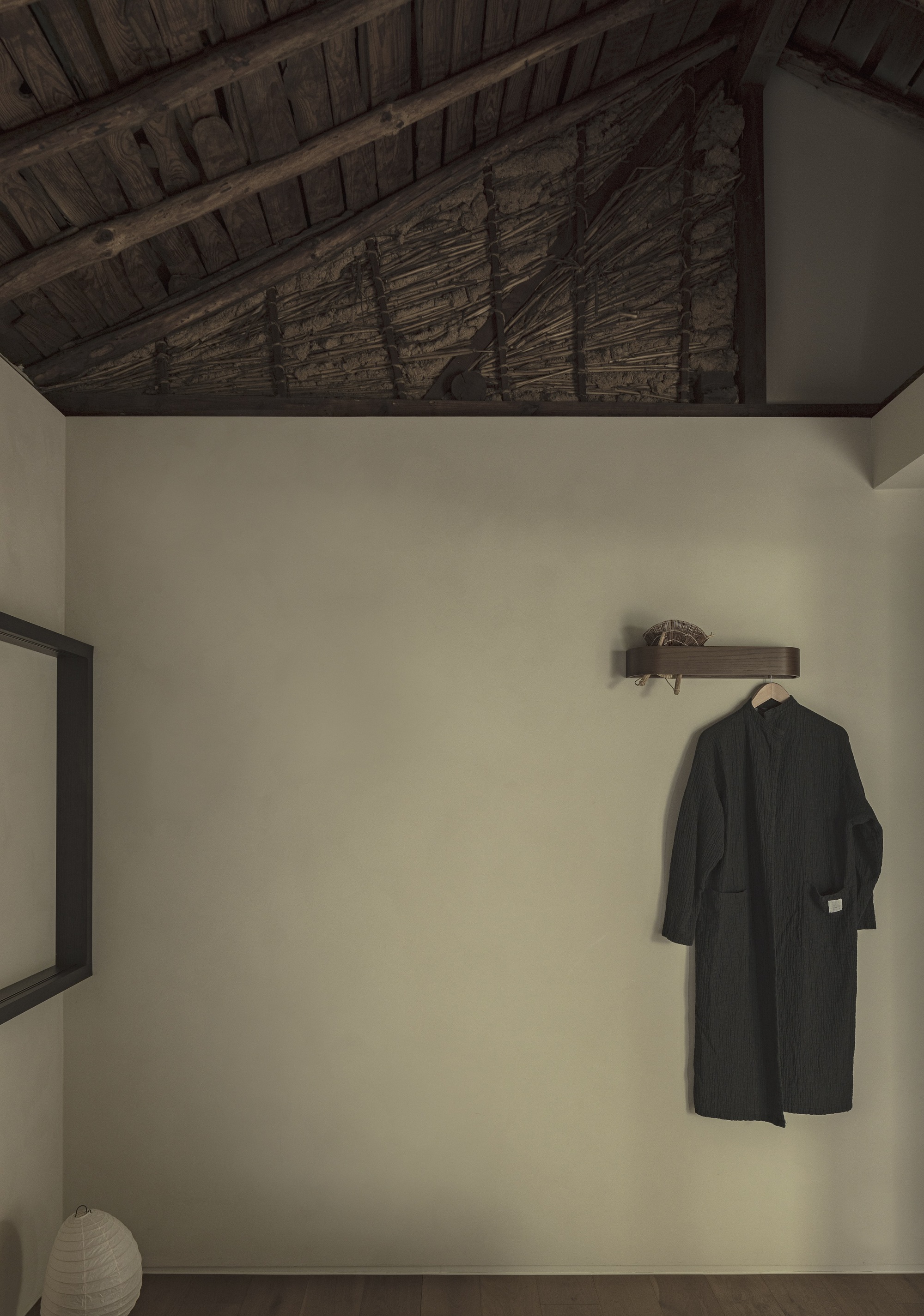 White plastered wall below exposed timber ceiling beams with a hanging coat and paper lantern