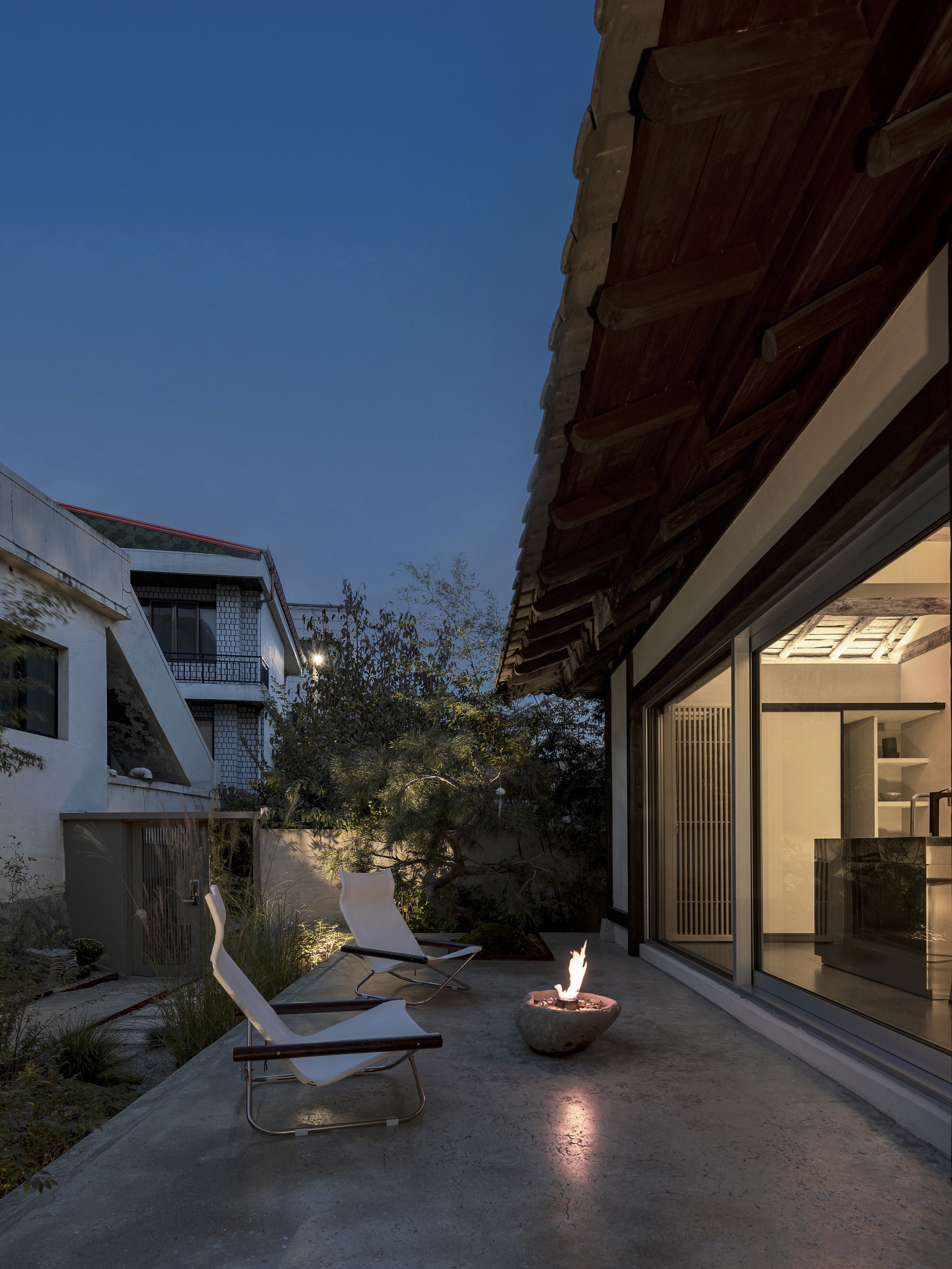 Courtyard terrace at dusk with fire bowl, lounge chairs and timber eaves above glazed walls