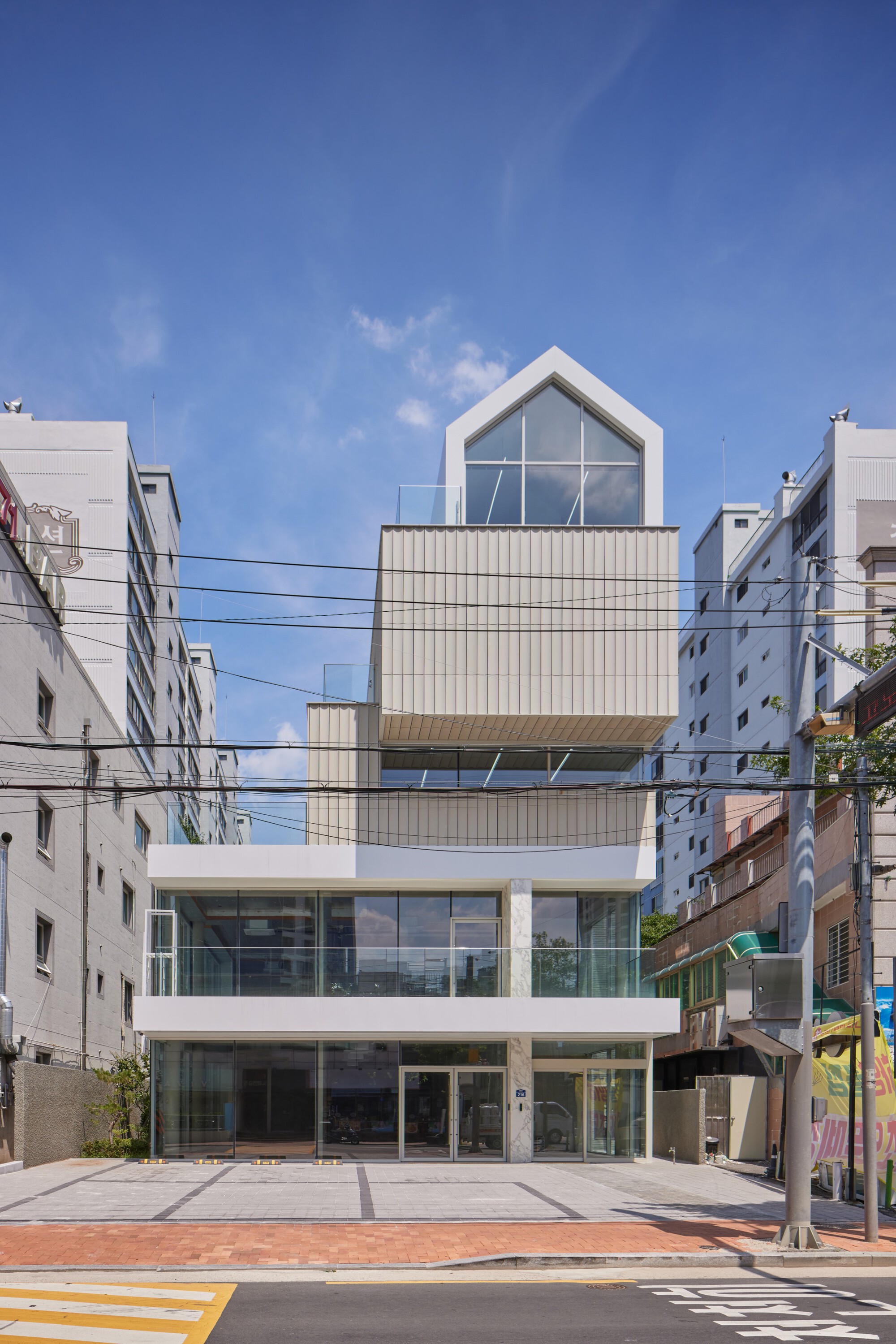 Front elevation showing gabled penthouse above cantilevered corrugated metal boxes and glass street level with overhead wires