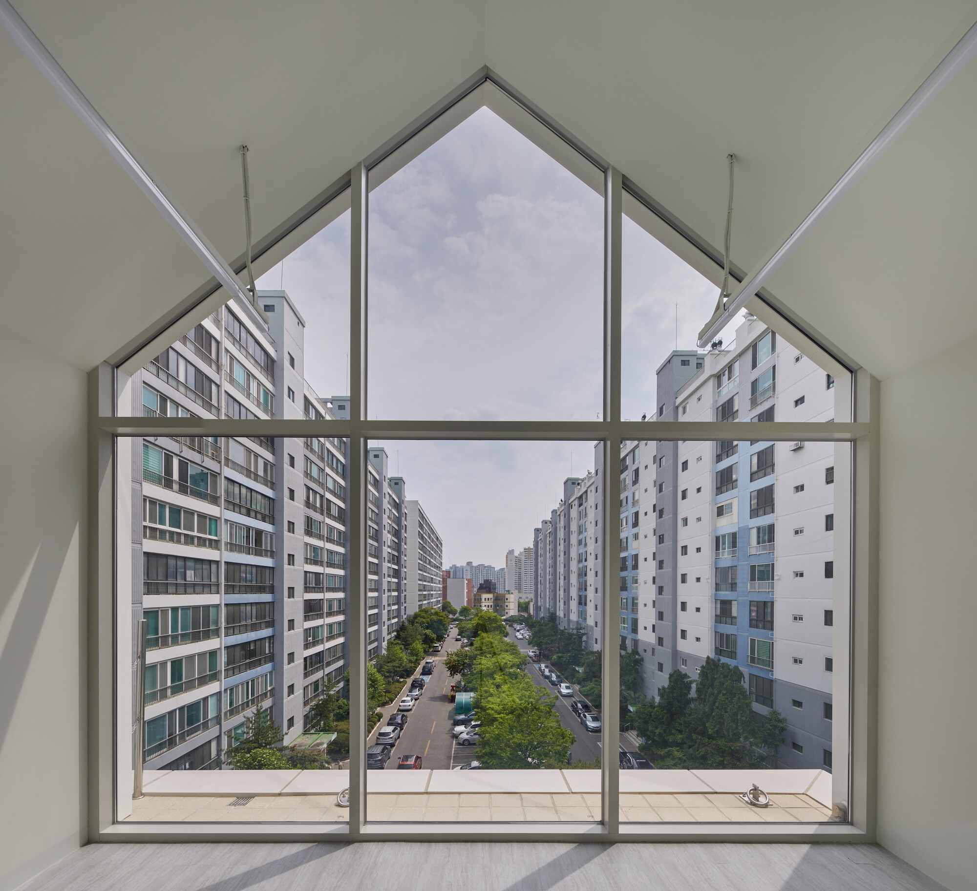 Interior view through gabled window framing tree-lined street and apartment blocks beyond
