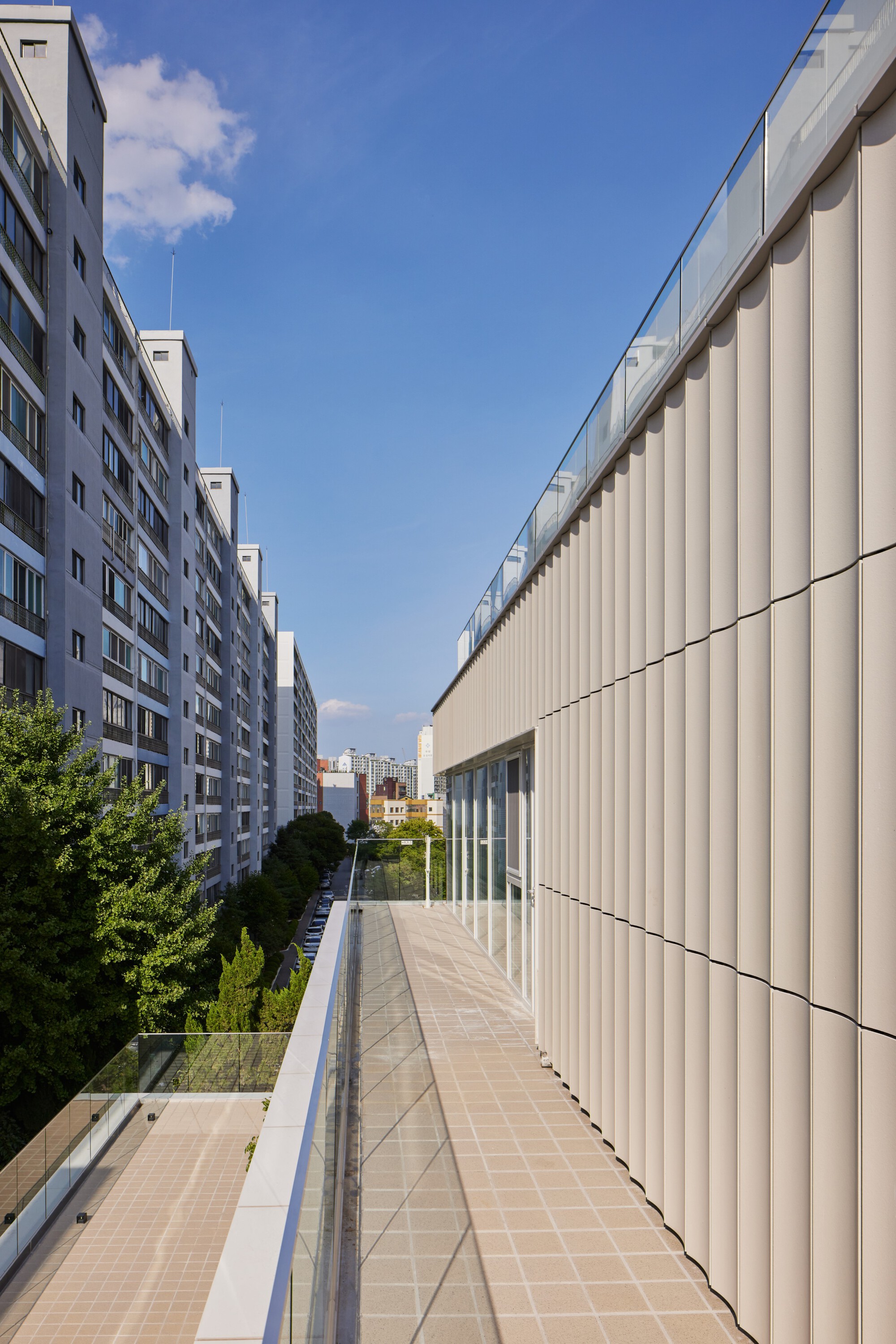 Upper terrace walkway with glass balustrade flanked by vertical metal facade and neighbouring apartment blocks