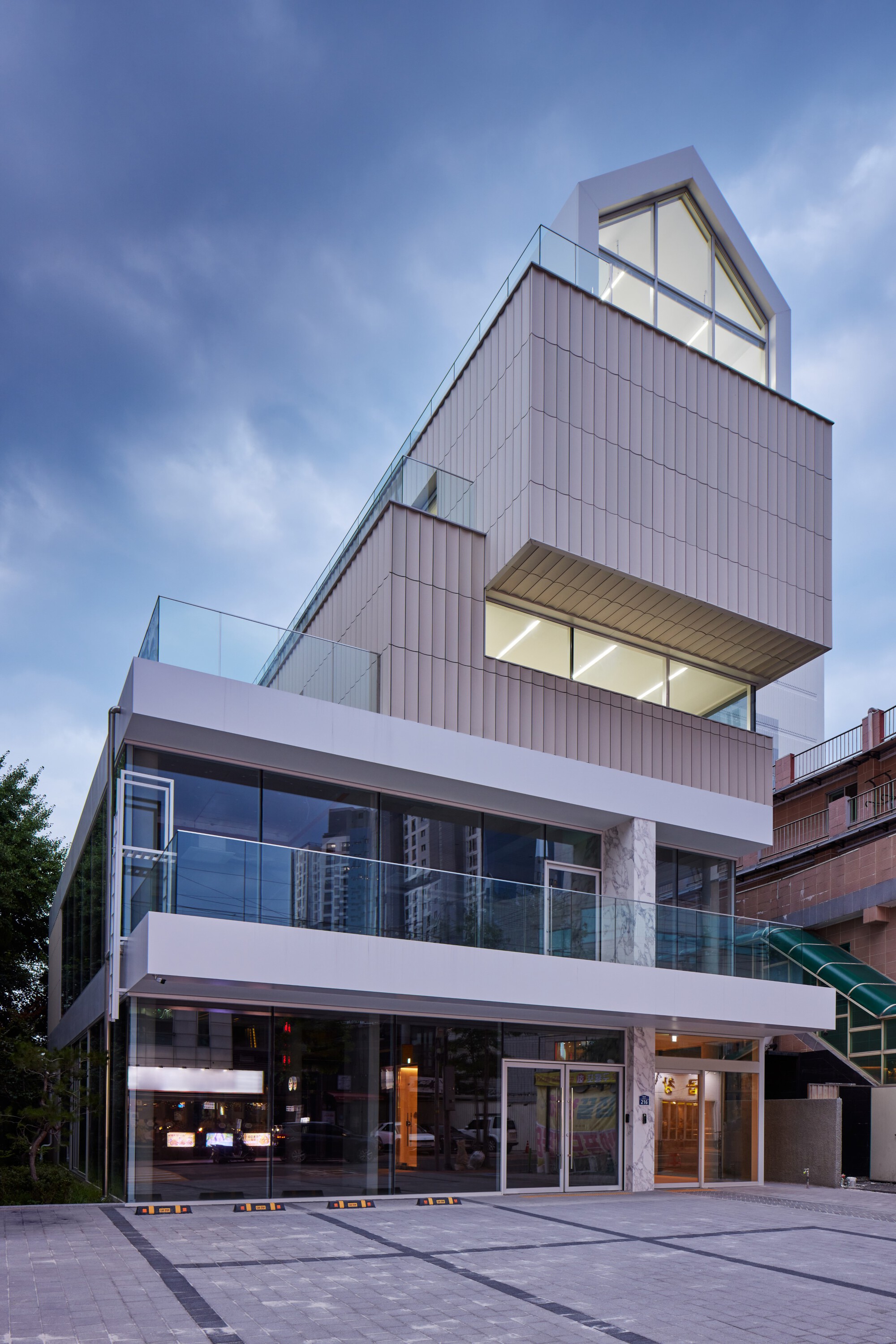 Street view showing stacked volumes with ribbed cladding, glass balconies, and ground-level storefront at twilight