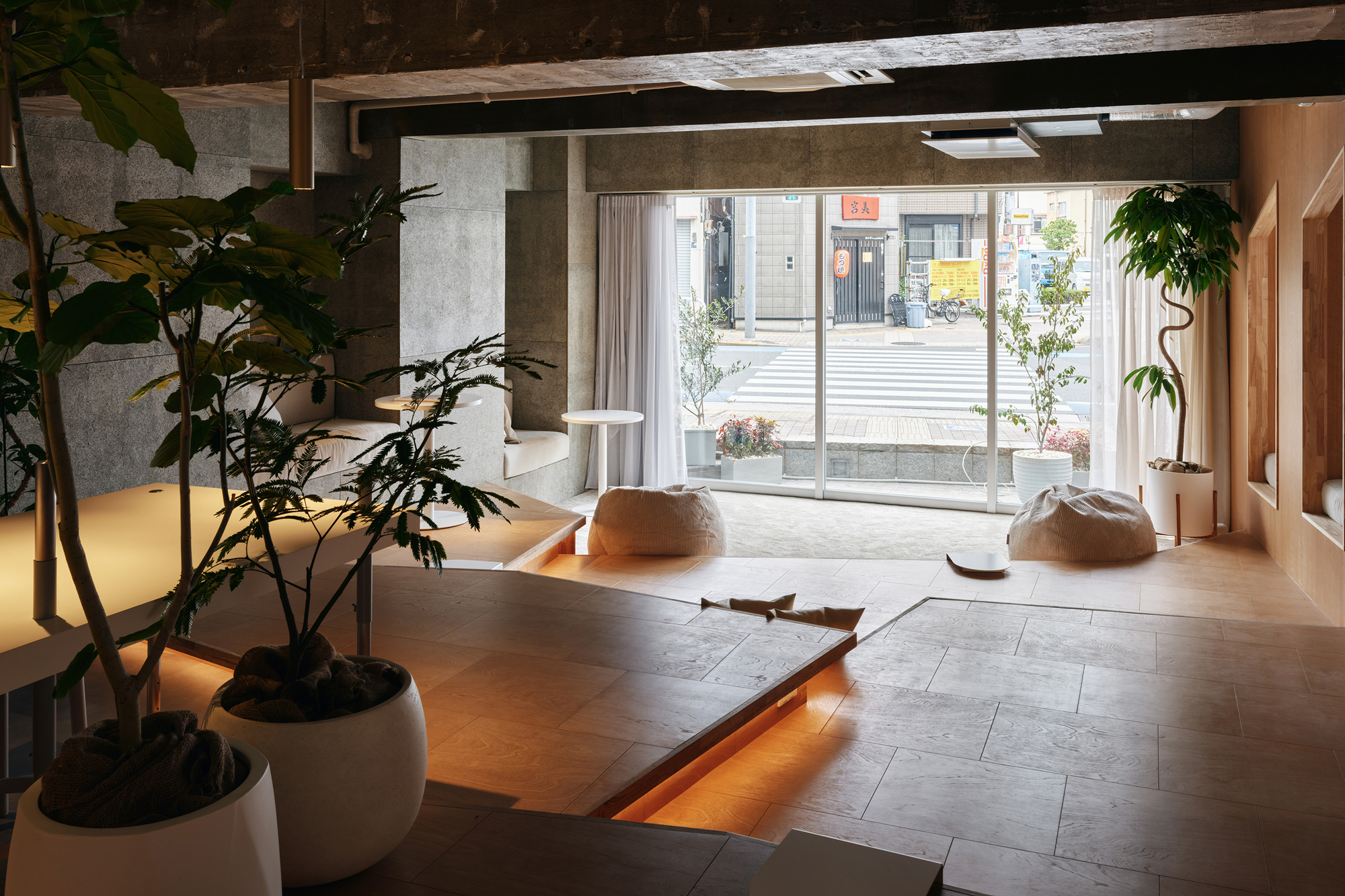 Interior lounge area with exposed concrete ceiling beams, potted plants, and a large glazed storefront opening to the street