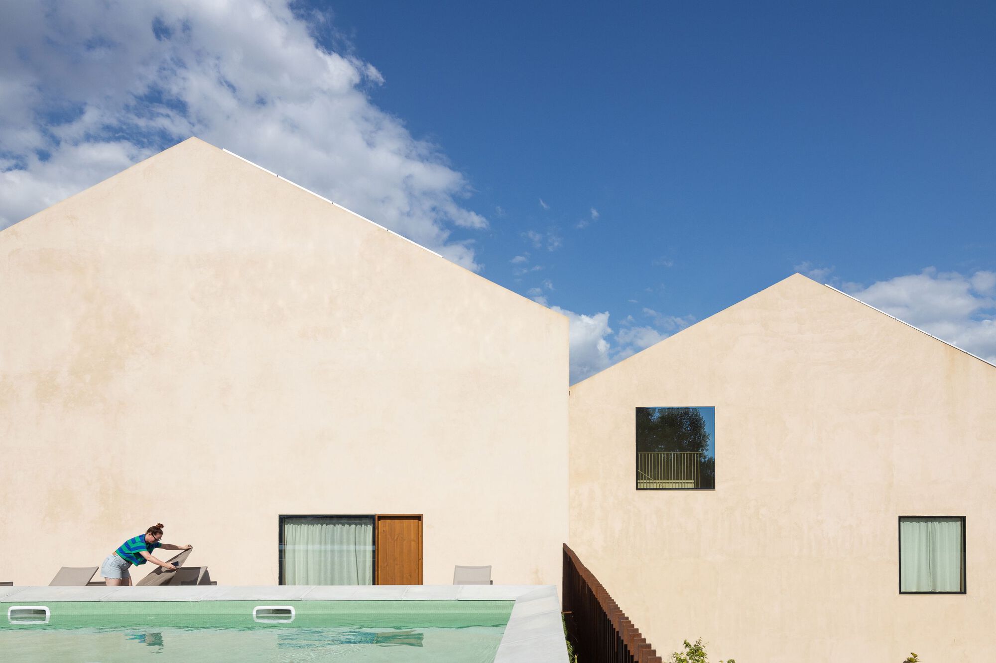 Two gabled volumes flanking a swimming pool with a cyclist passing under afternoon clouds