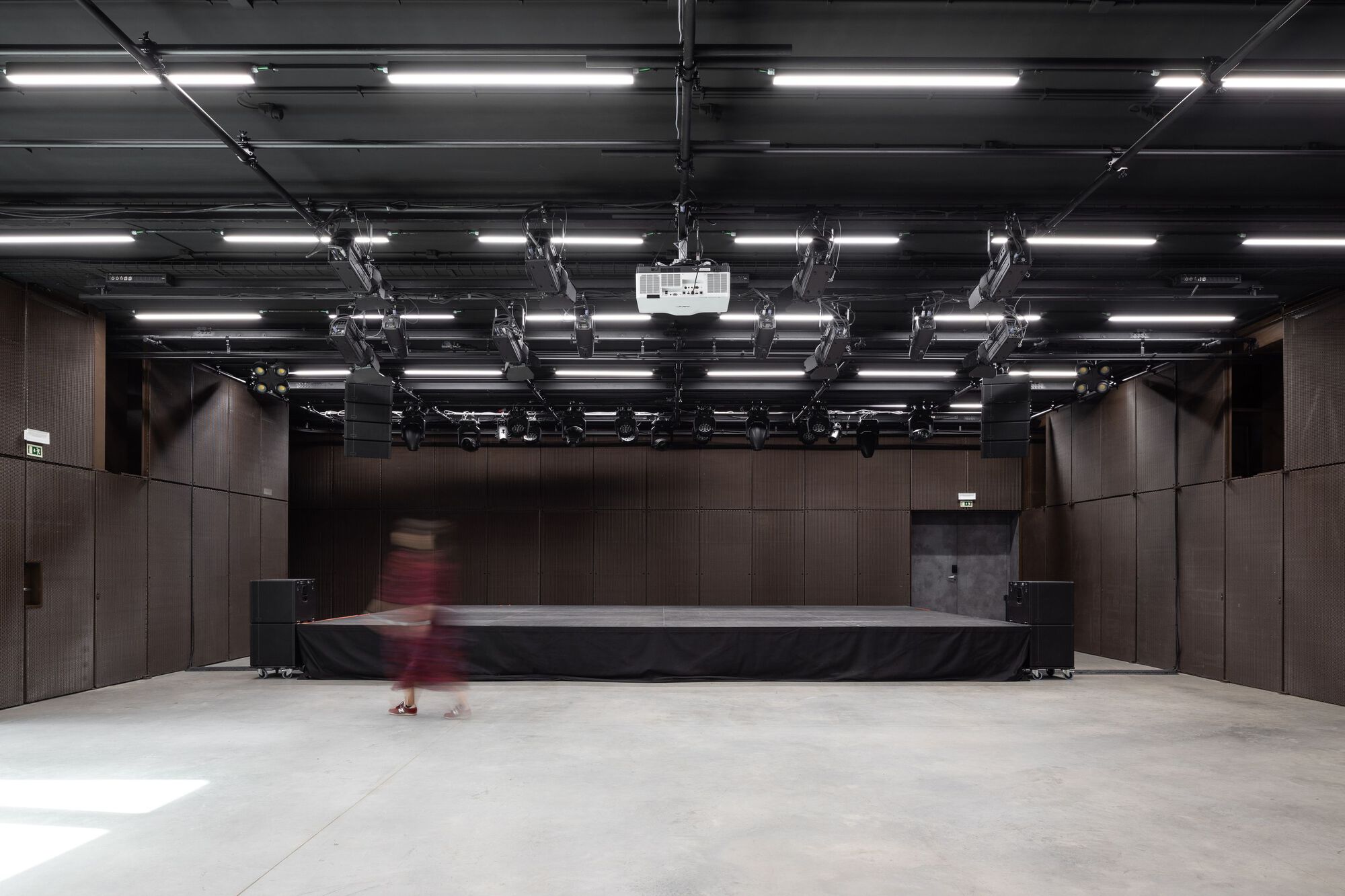 Performance hall with exposed ceiling lighting grid, dark timber wall panels, and a figure in motion