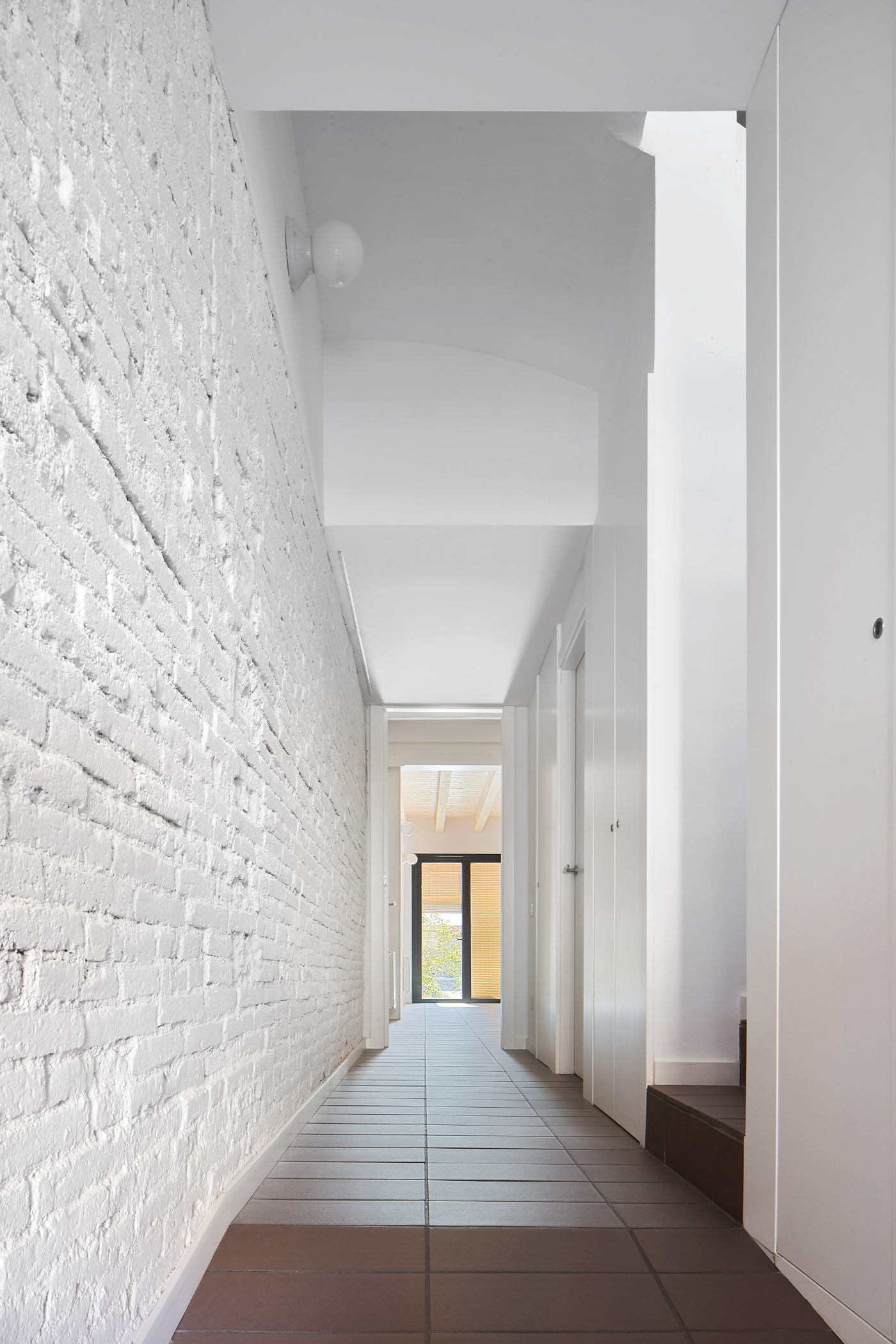 Narrow corridor with painted white brick wall and terracotta tile floor leading to a distant window