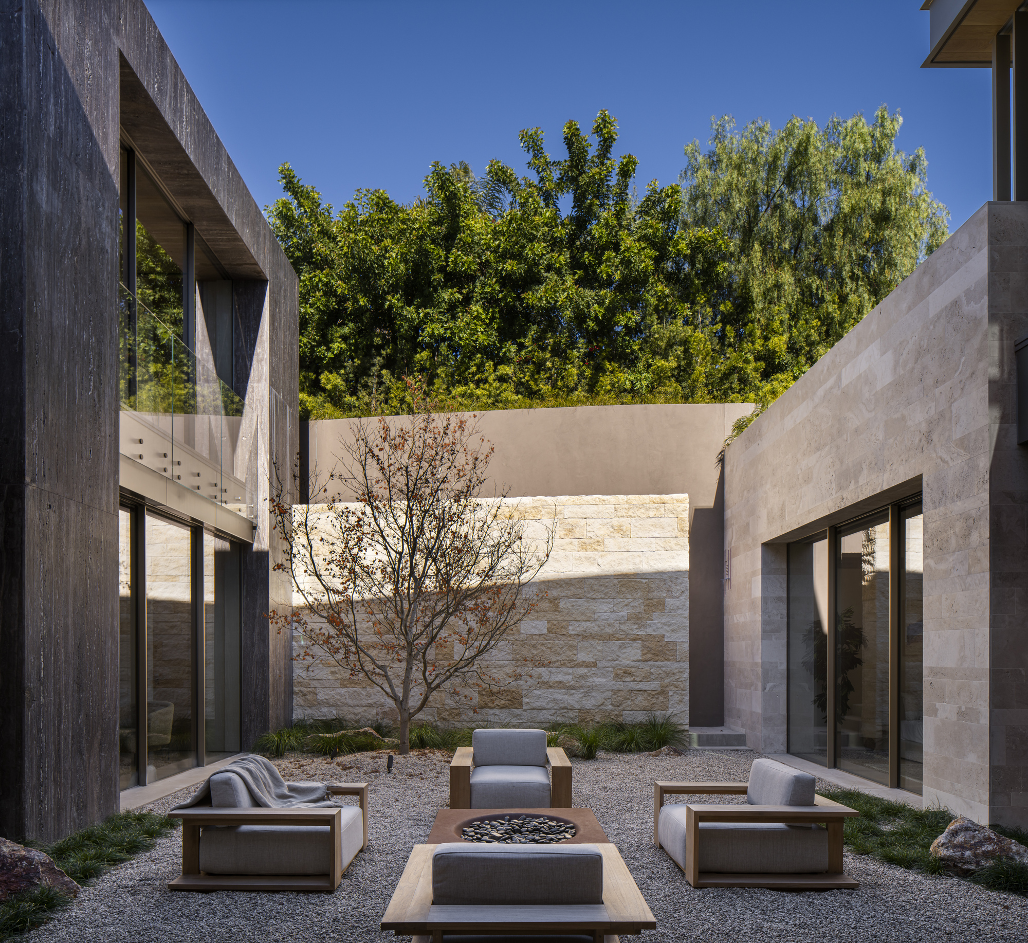 Courtyard with gravel ground plane, central fire pit, single tree, and stone walls beneath overhanging greenery