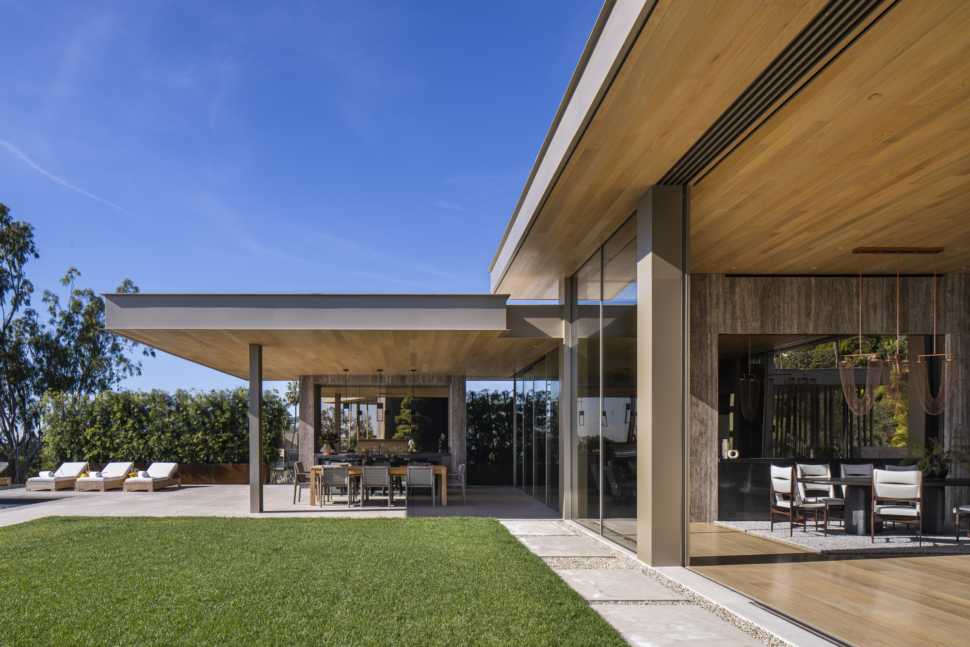 Covered terrace with timber ceiling and steel columns opening to a lawn under clear blue sky