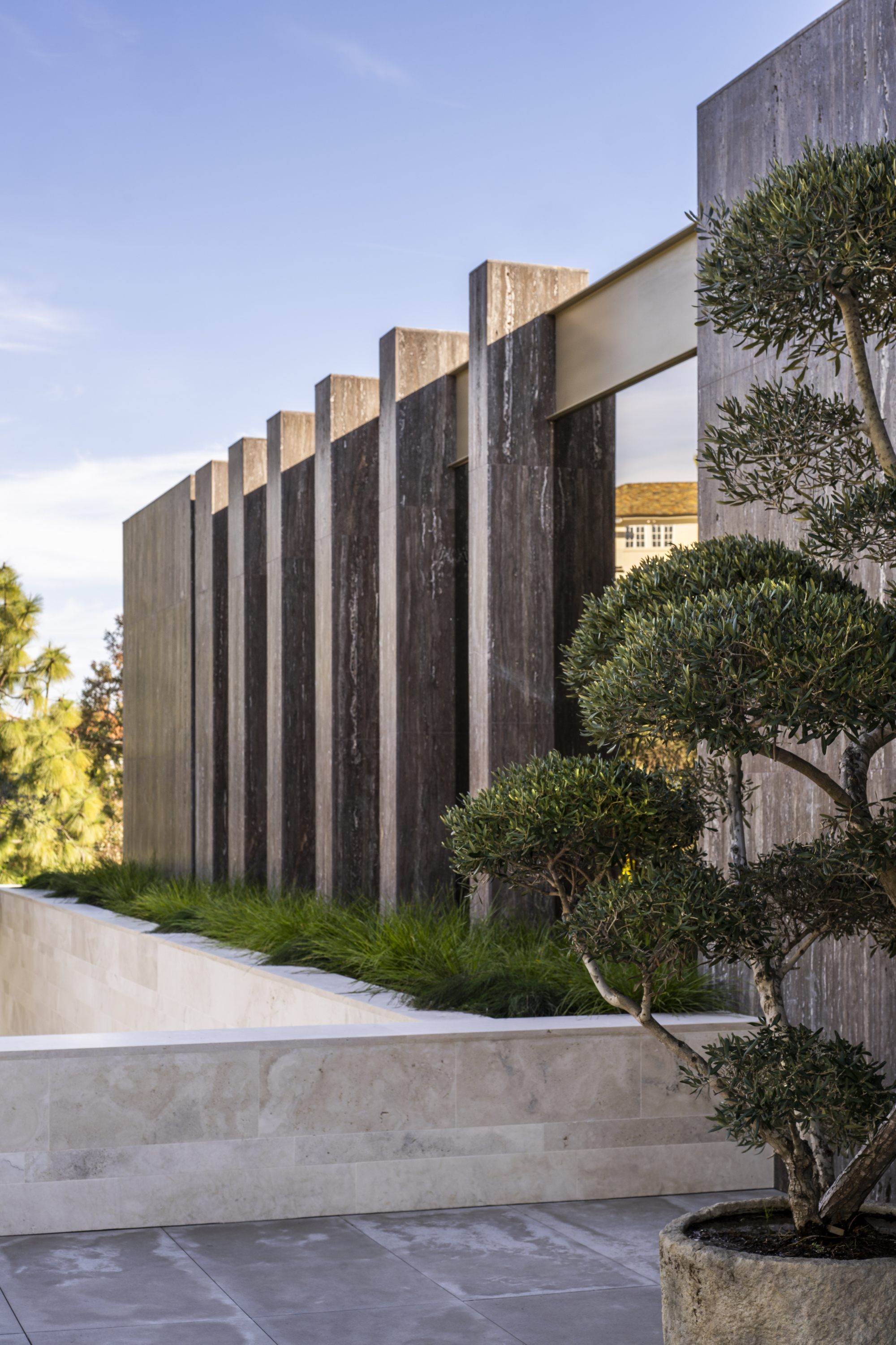 Vertical concrete fin screen beside stone entry steps and potted olive tree at dusk