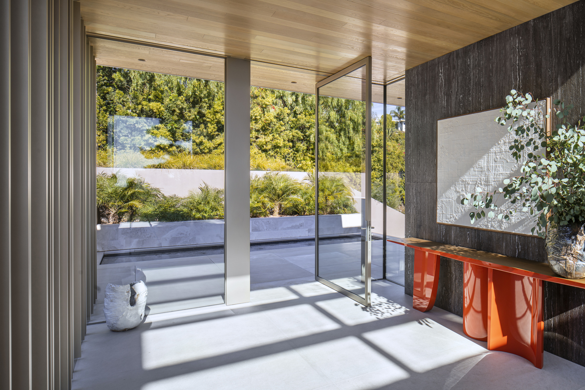 Entry foyer with glass pivot door, orange lacquered console, and view to planted terrace in sunlight