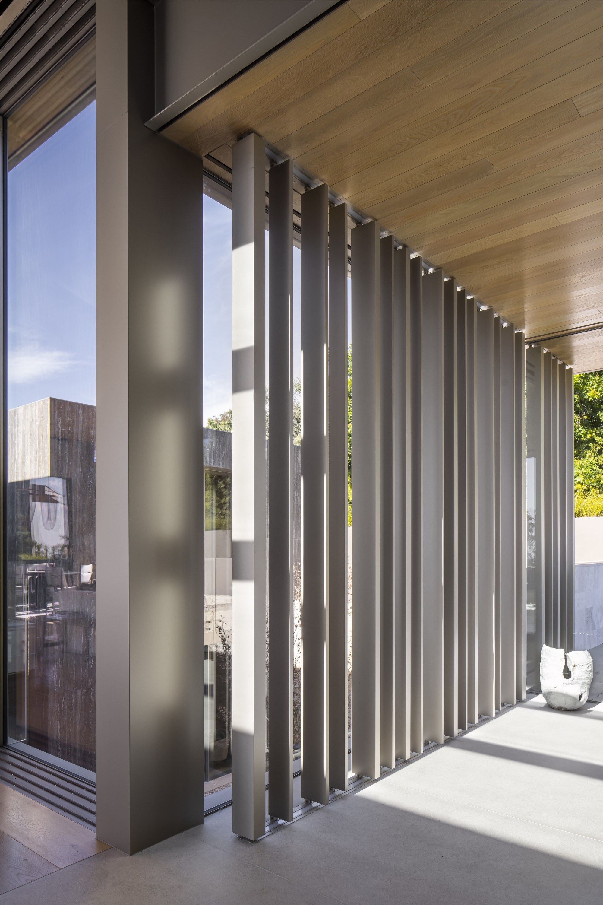 Exterior terrace framed by vertical metal louvers beneath a timber ceiling in bright sunlight