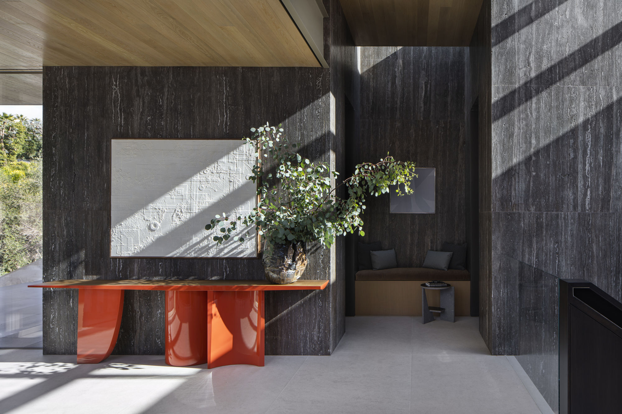 Interior view of dark vertical timber walls with an orange sculptural console table and morning sunlight streaming through openings