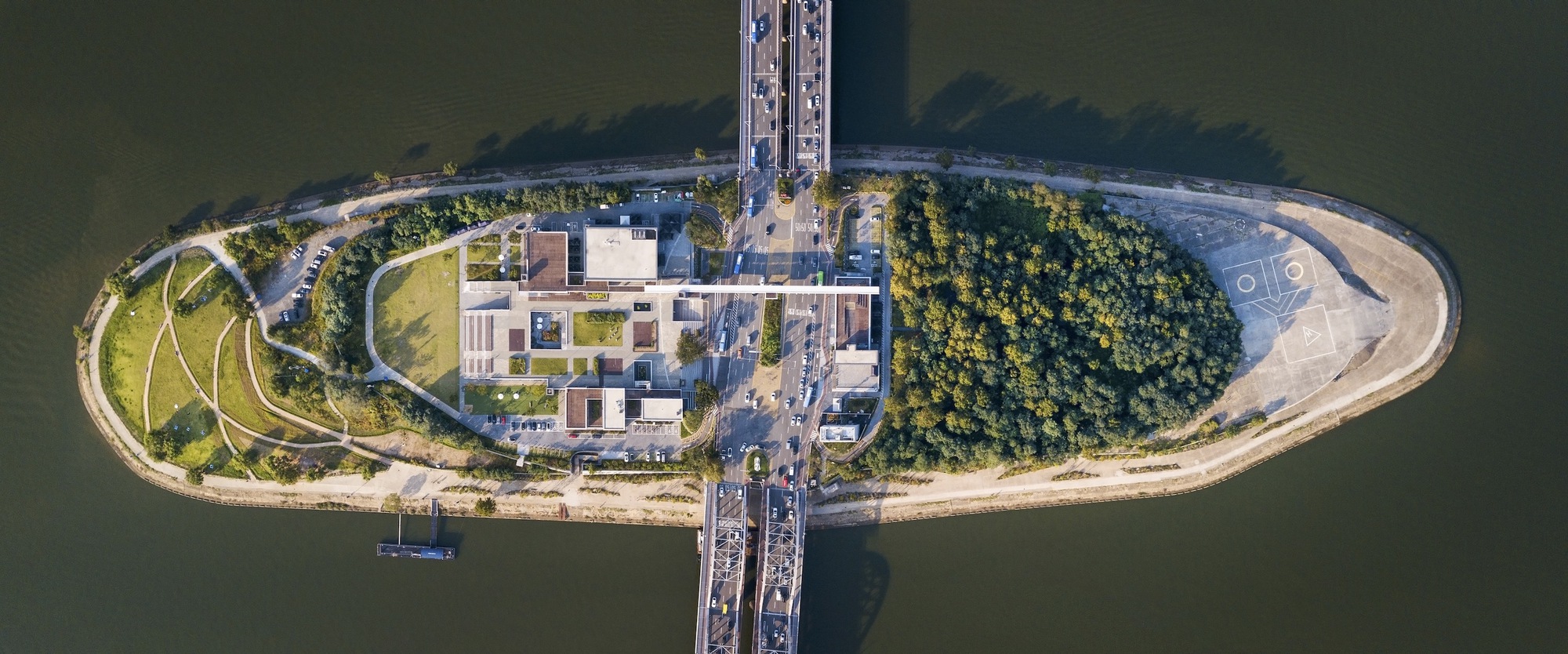 Top-down view of the oval island showing low-rise volumes surrounded by planted areas and bridge connections