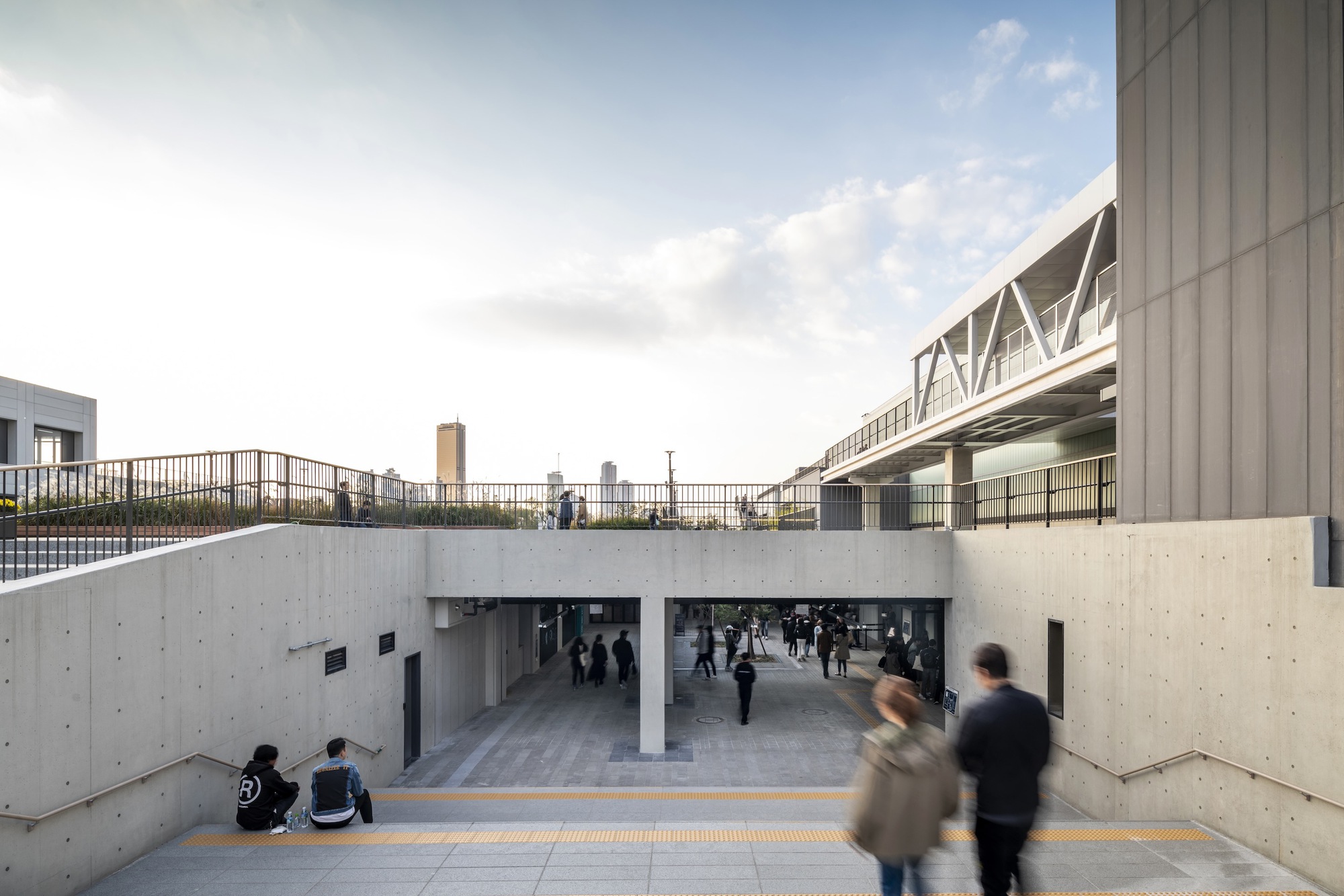 Concrete sunken passageway with pedestrians moving through beneath an elevated walkway