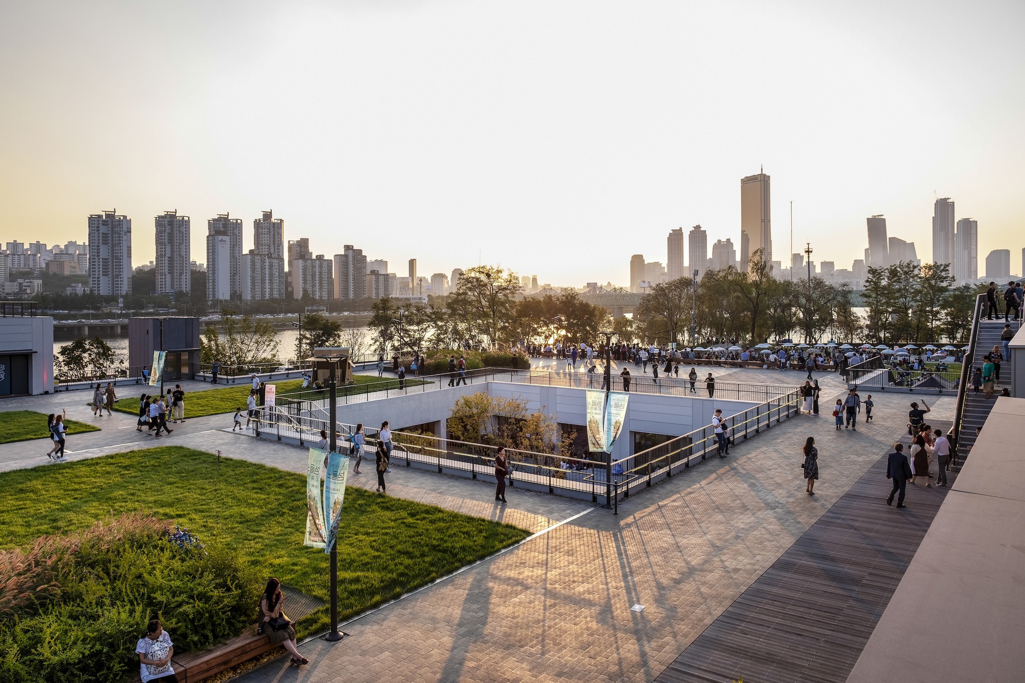 Waterfront park with lawns and sunken plaza at dusk with distant skyline reflected in the river