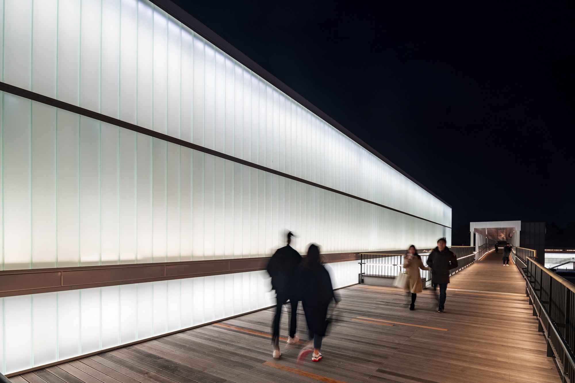 Elevated timber walkway along a backlit translucent panel wall with blurred figures at night