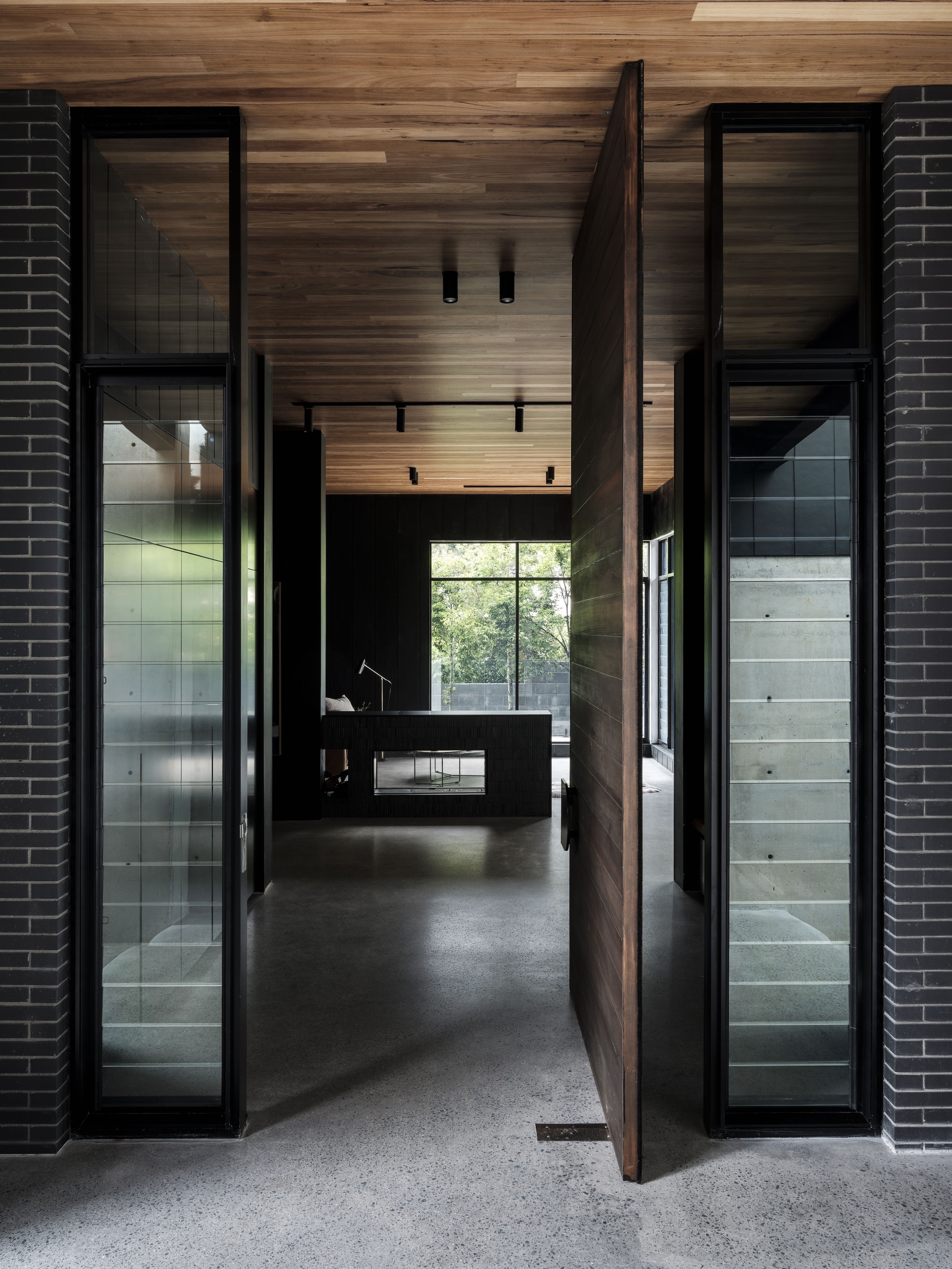 Entry hallway with timber-clad ceiling and pivot door flanked by glass panels with dark brick walls