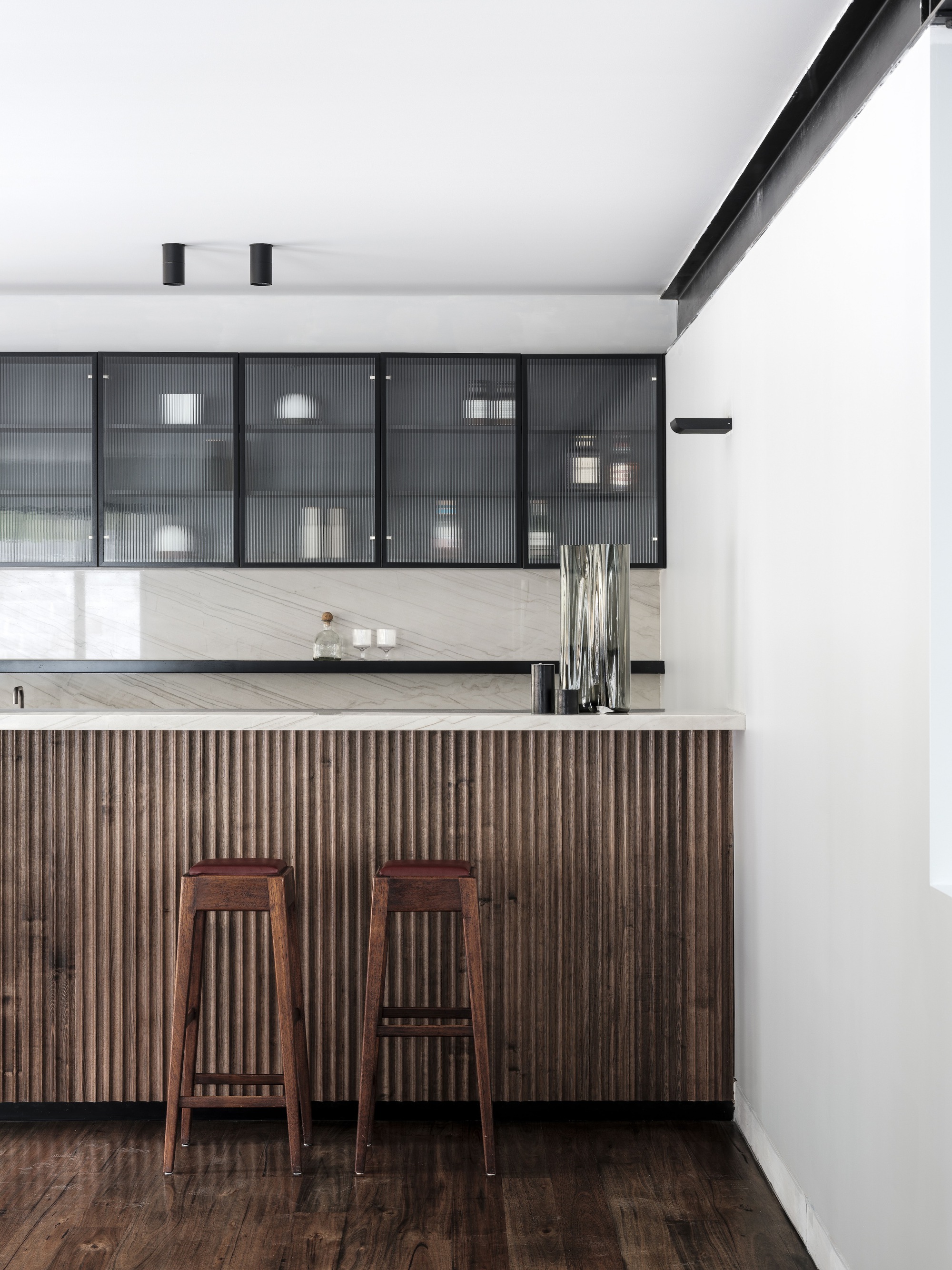 Kitchen island with vertical timber slat cladding and two wooden stools beneath reeded glass cabinets