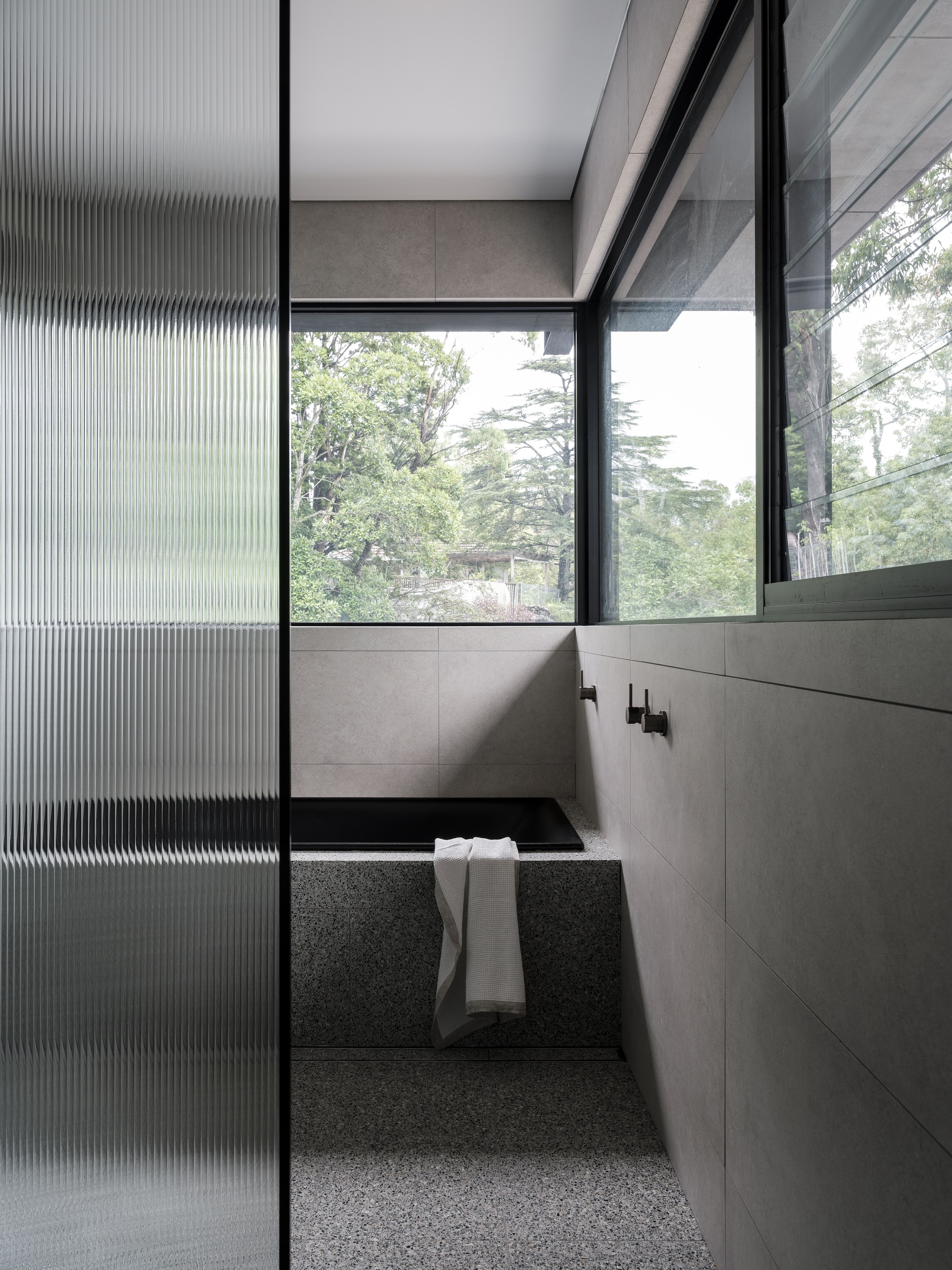 Sunken bathtub with terrazzo surround framed by fluted glass screen and windows overlooking garden