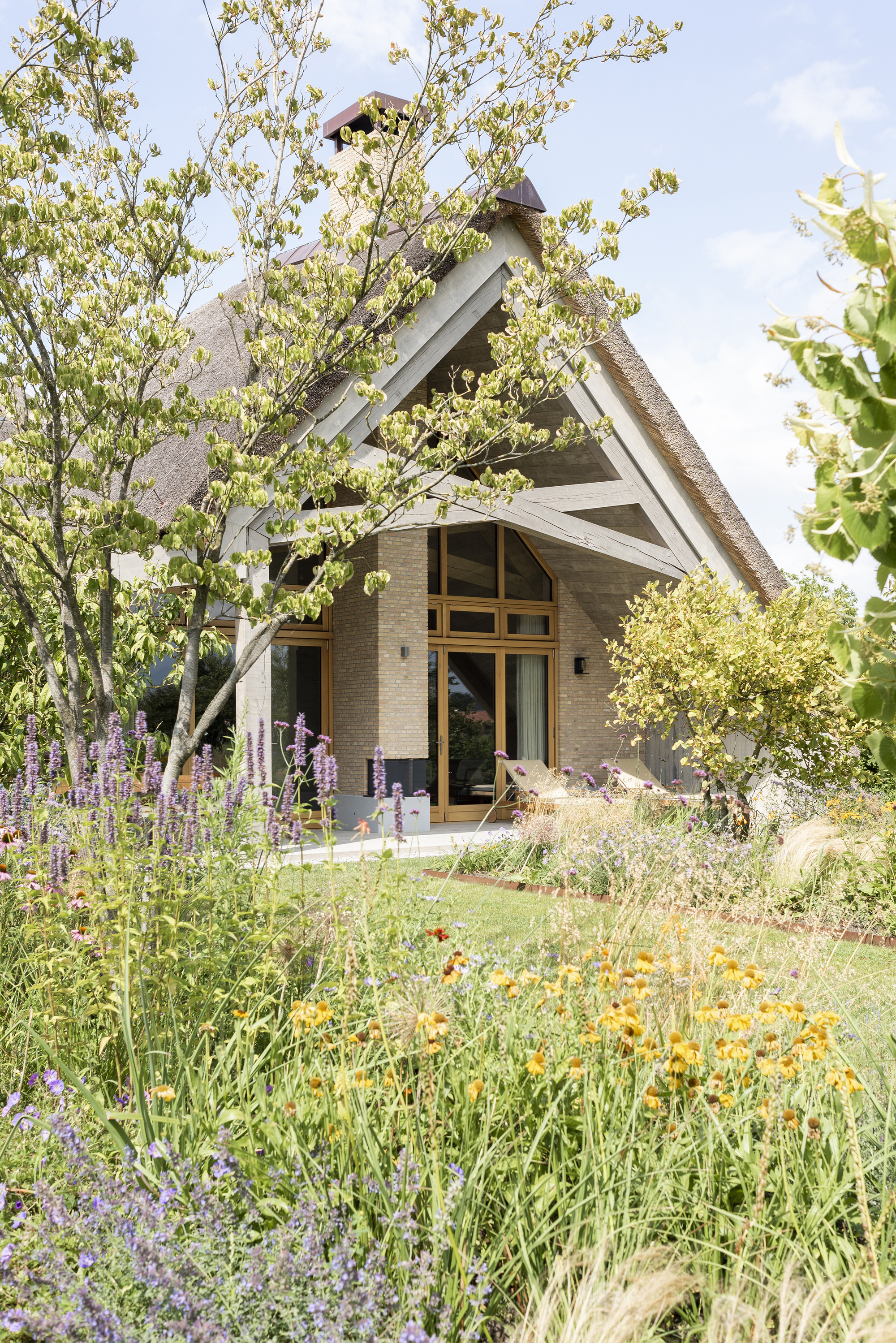 Entrance facade with thatched gable roof surrounded by flowering perennials and young trees