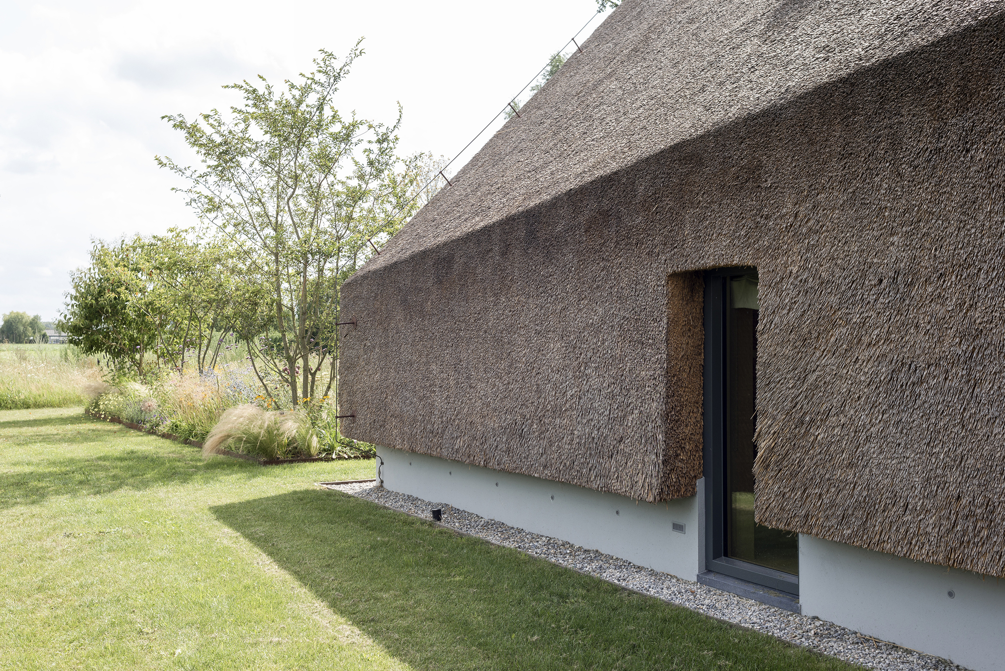 Thatched roof extending down to a white plinth with a narrow glazed door and gravel path