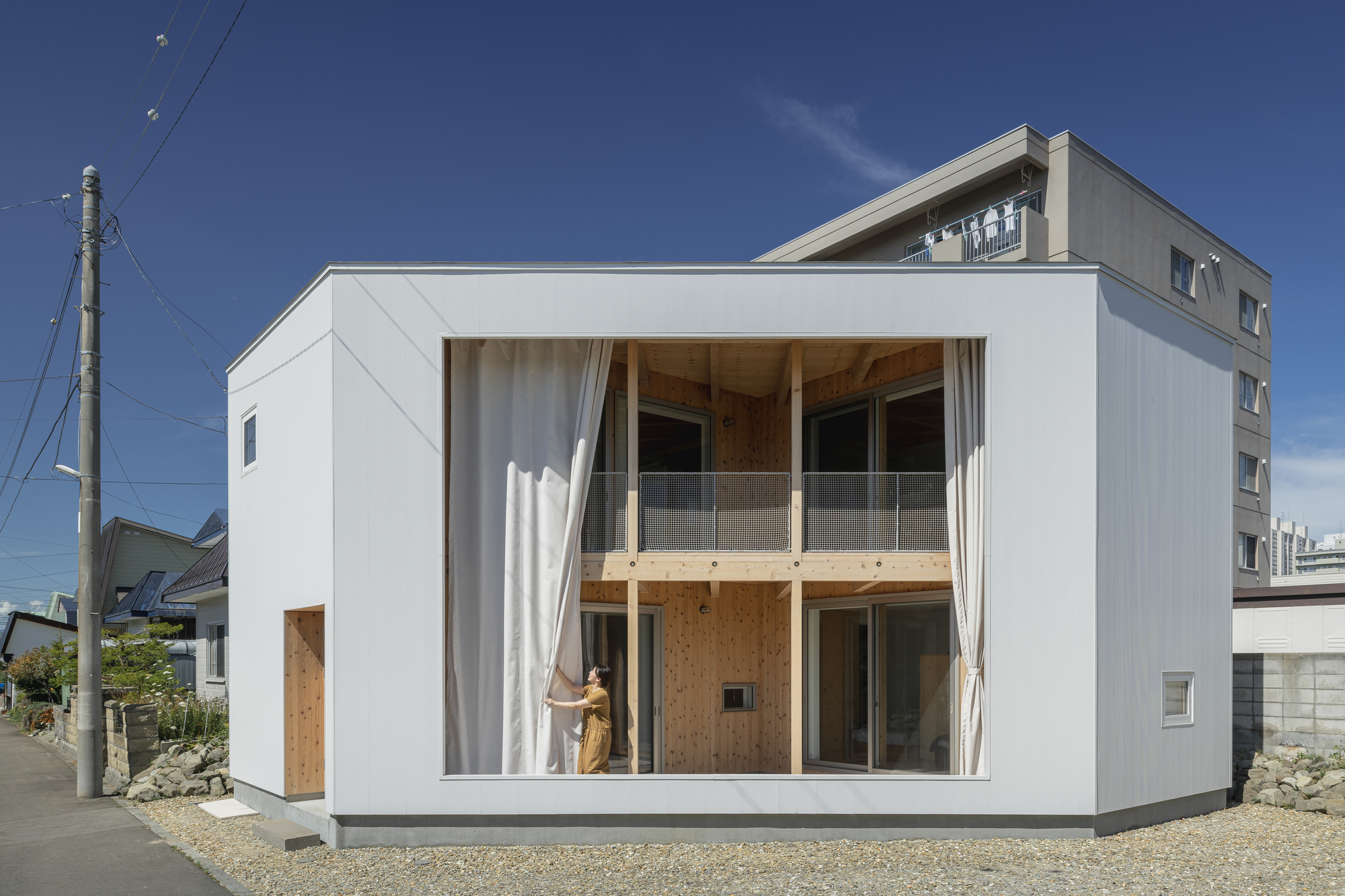White box facade with floor-to-ceiling glazing and sheer curtains pulled back by a figure inside
