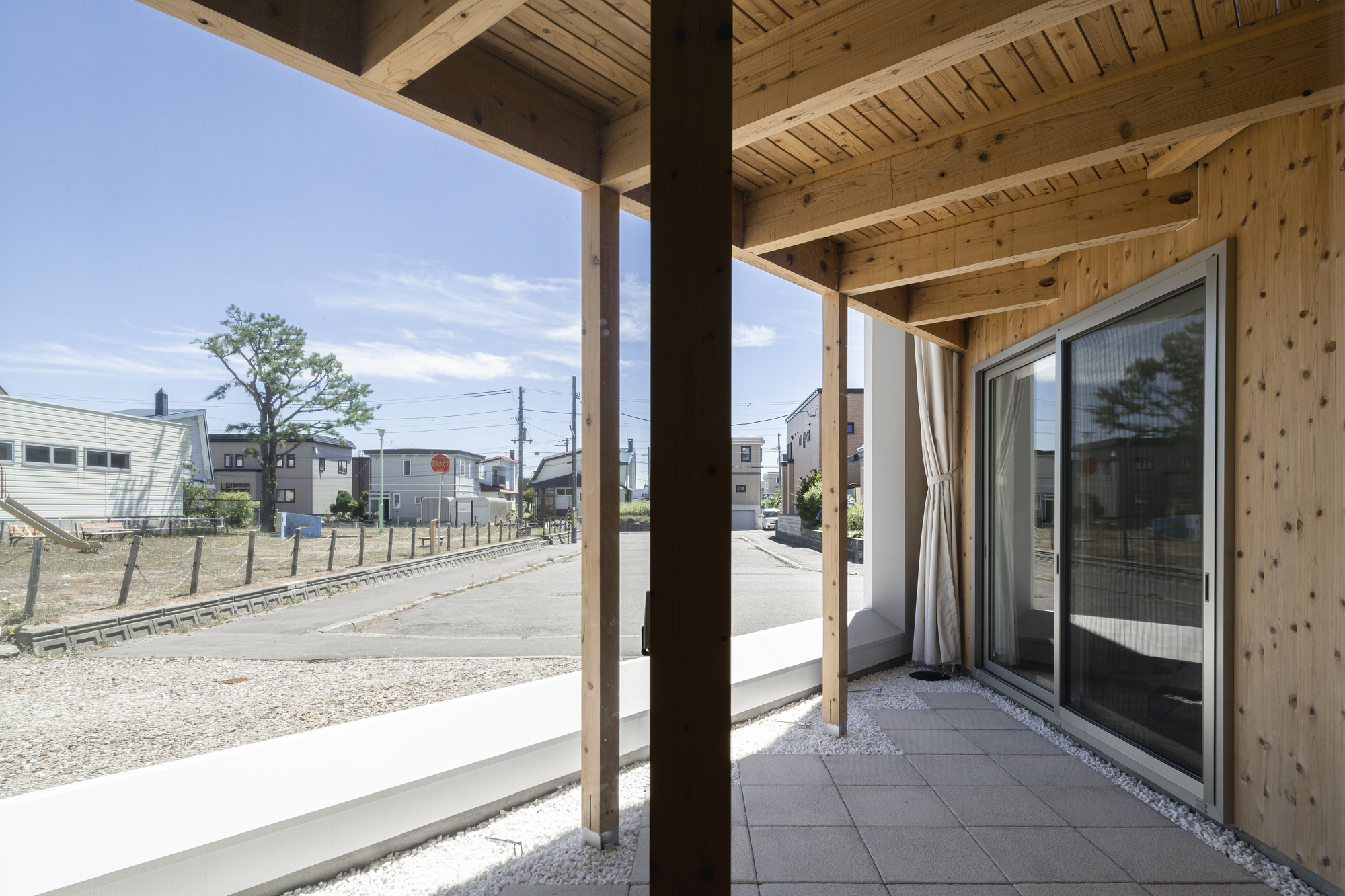 Covered terrace with timber posts and joists overlooking the gravel lot and neighboring houses