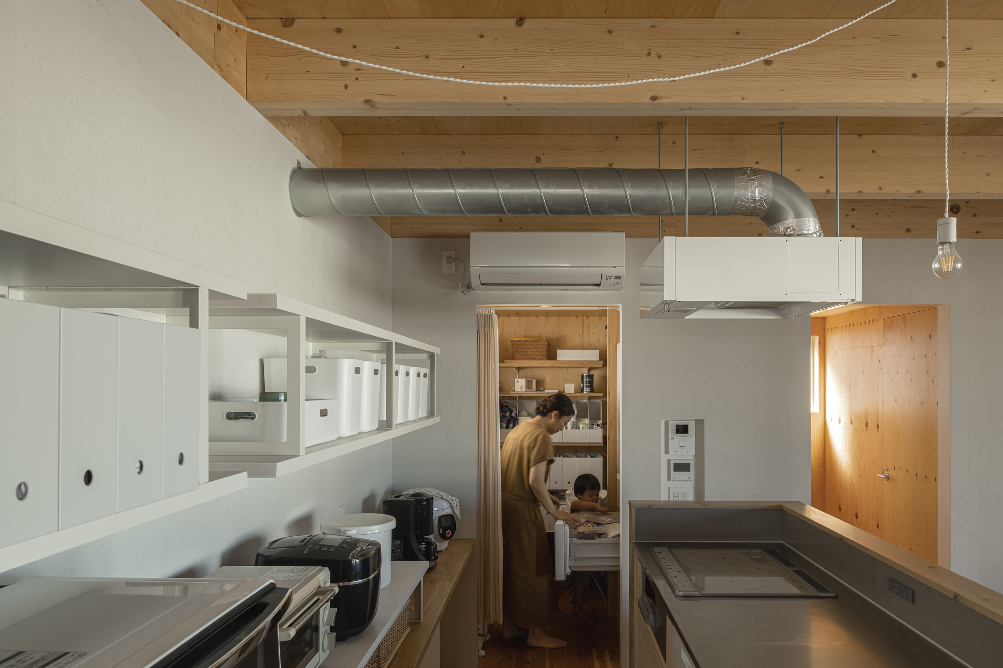 Kitchen workspace with overhead exposed duct, white storage boxes, and figures visible through pantry doorway