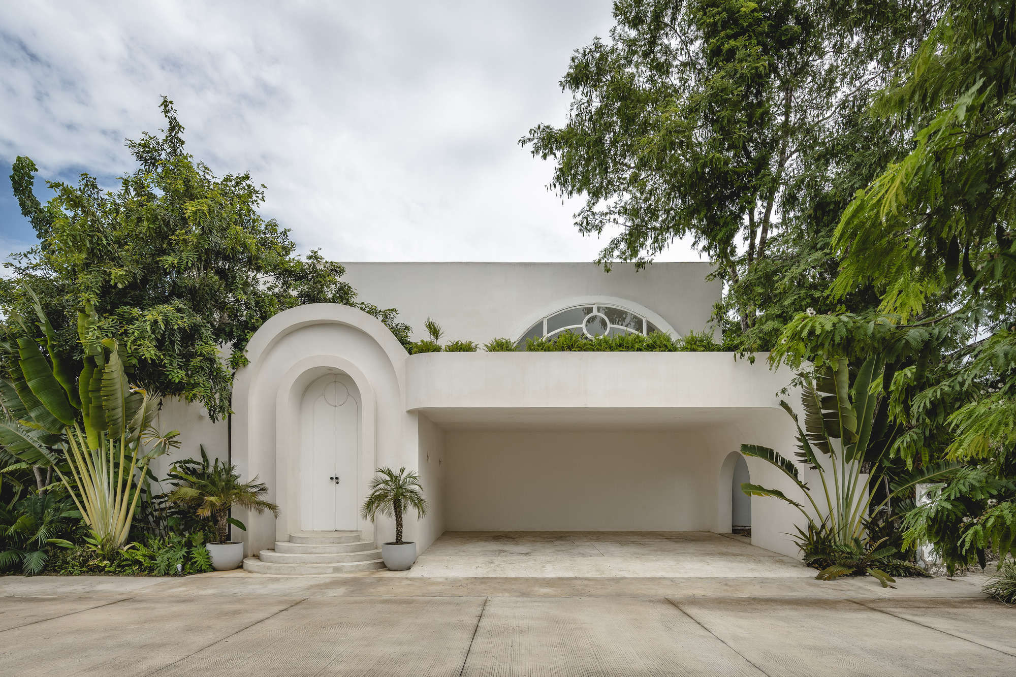 White facade with arched entrance, recessed garage, and circular window framed by banana plants and tropical trees