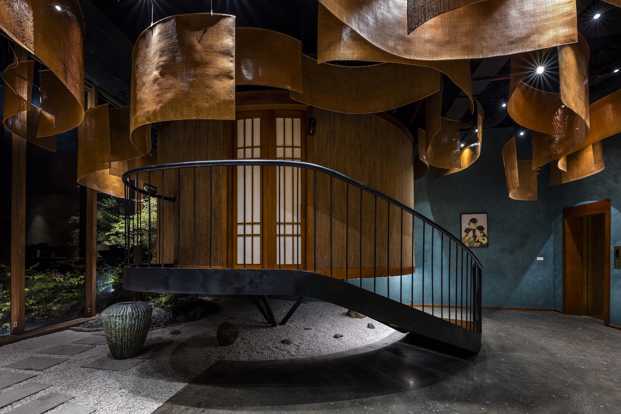 Curved staircase with black steel railings rising from a raked gravel garden bed beneath woven copper ceiling panels