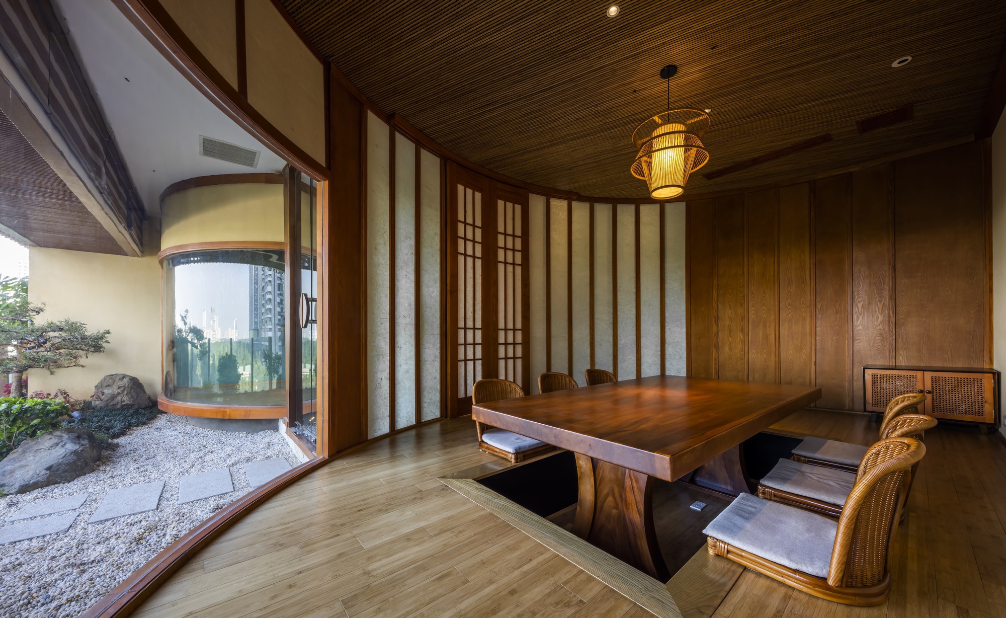 Timber dining table beside curved glass walls overlooking a small rock garden with raked gravel