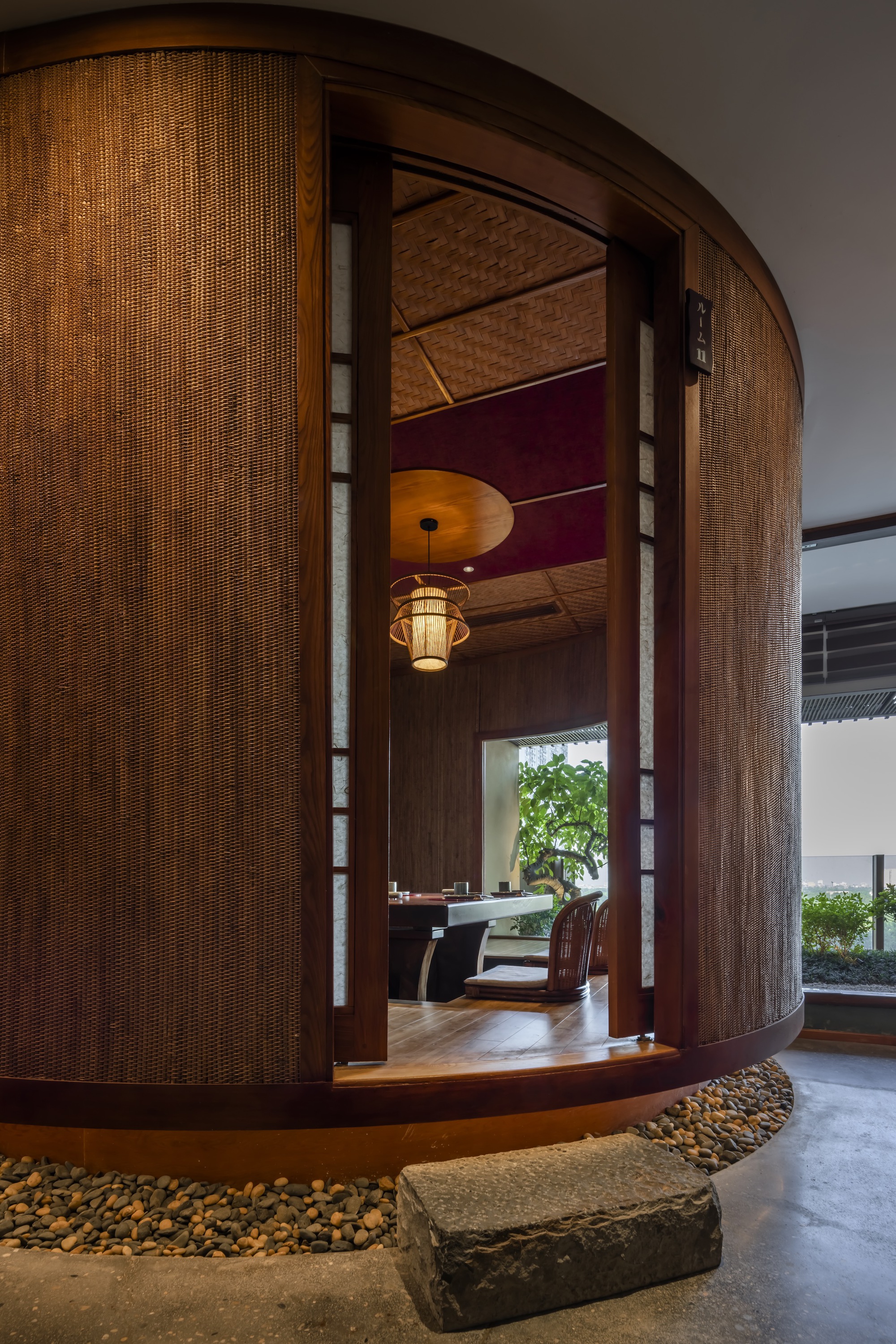 Curved woven rattan entry portal with stone steps surrounded by river pebbles under warm interior lighting