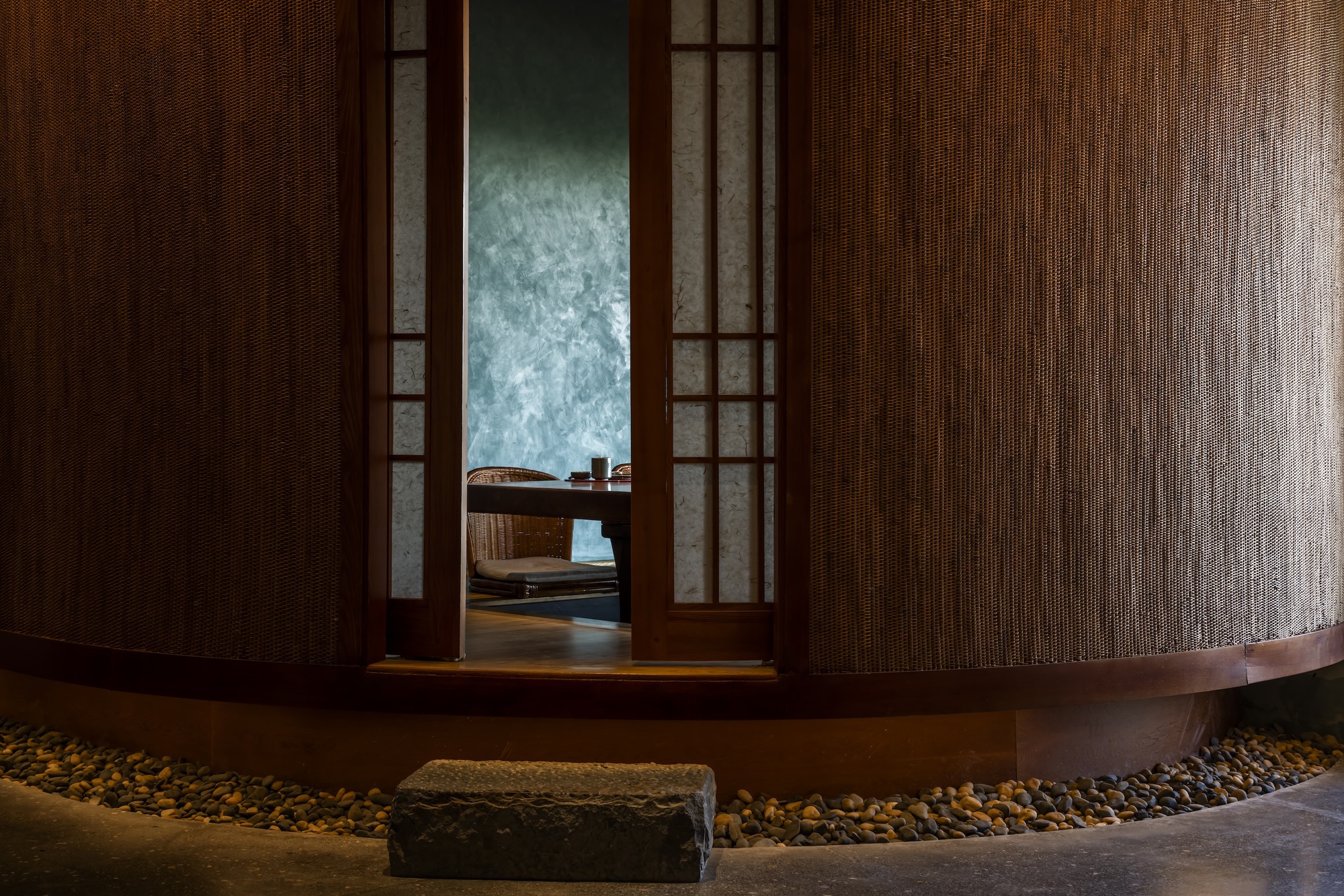 Narrow timber and translucent panel doorway framed by woven rattan walls with stone steps at base