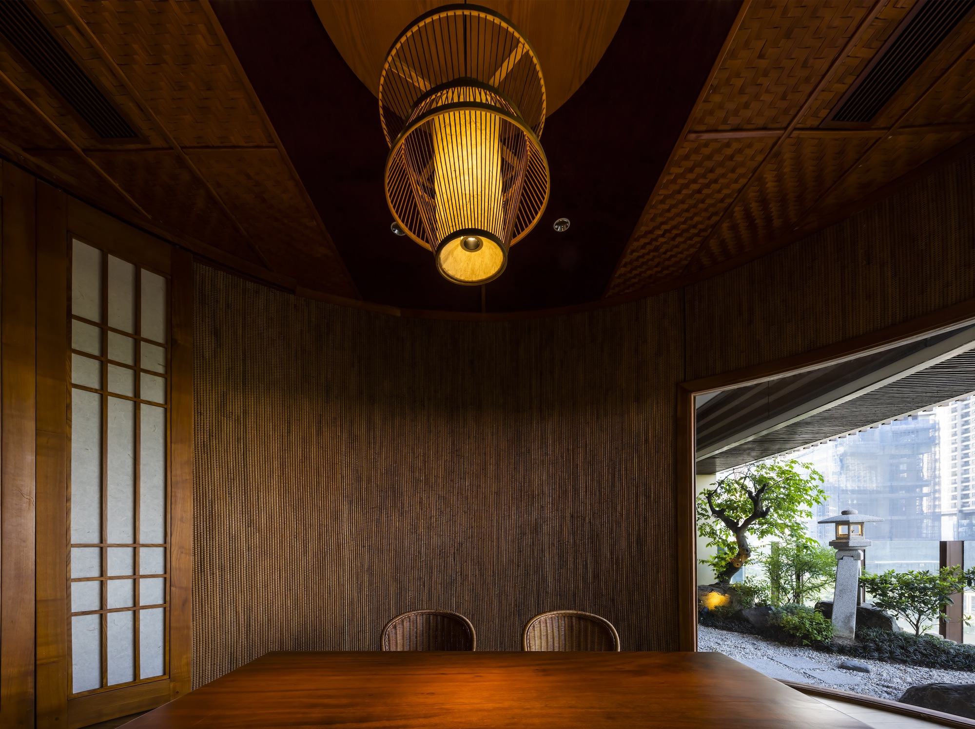Private dining room with textured woven wall panels, bamboo lantern and opening to planted courtyard beyond