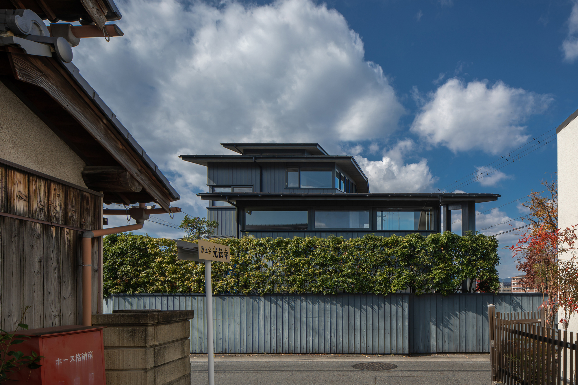 Street view of the layered volumes with deep overhangs rising above a hedge-lined corrugated metal fence
