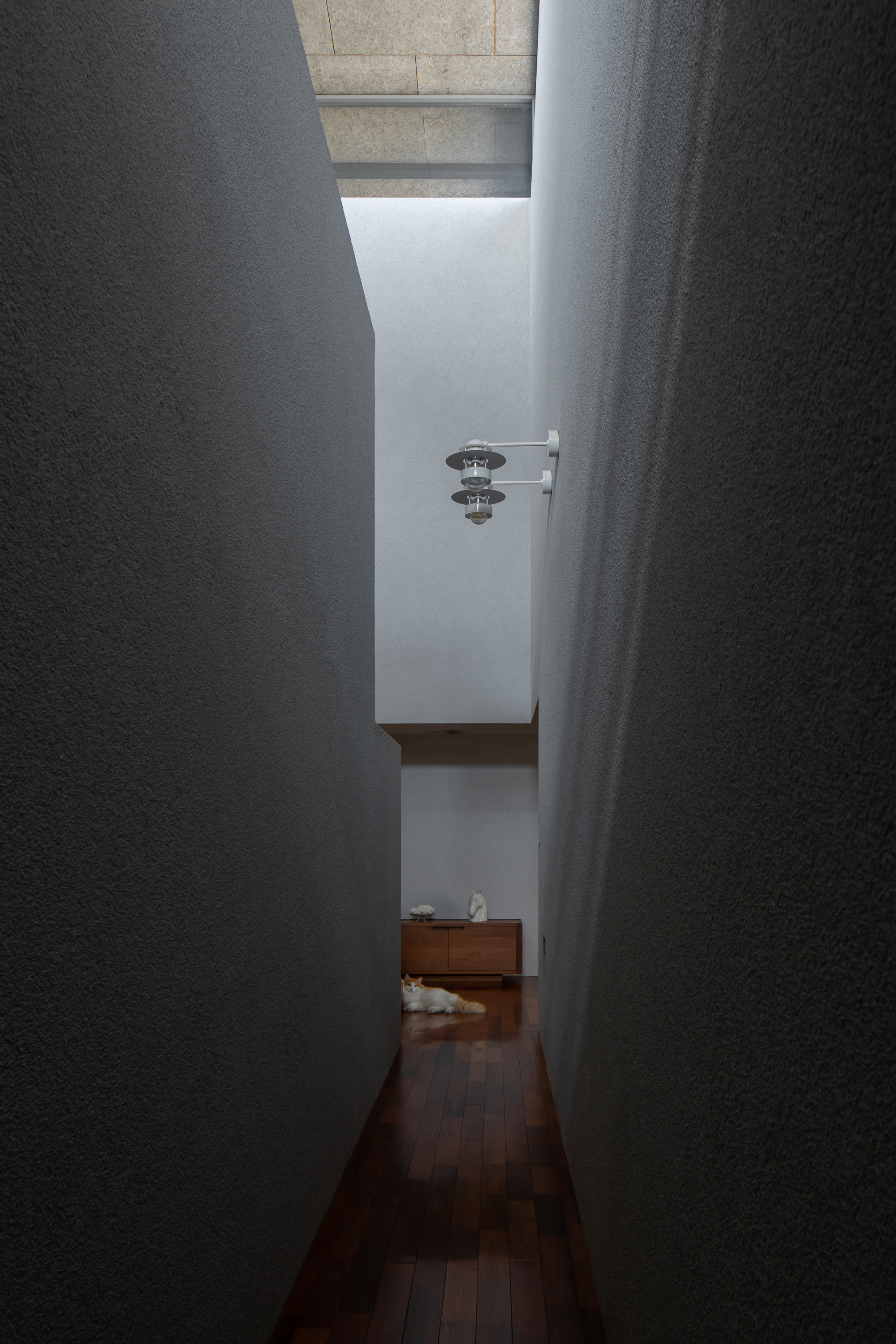 Narrow corridor with textured plaster walls opening to a skylit space beyond