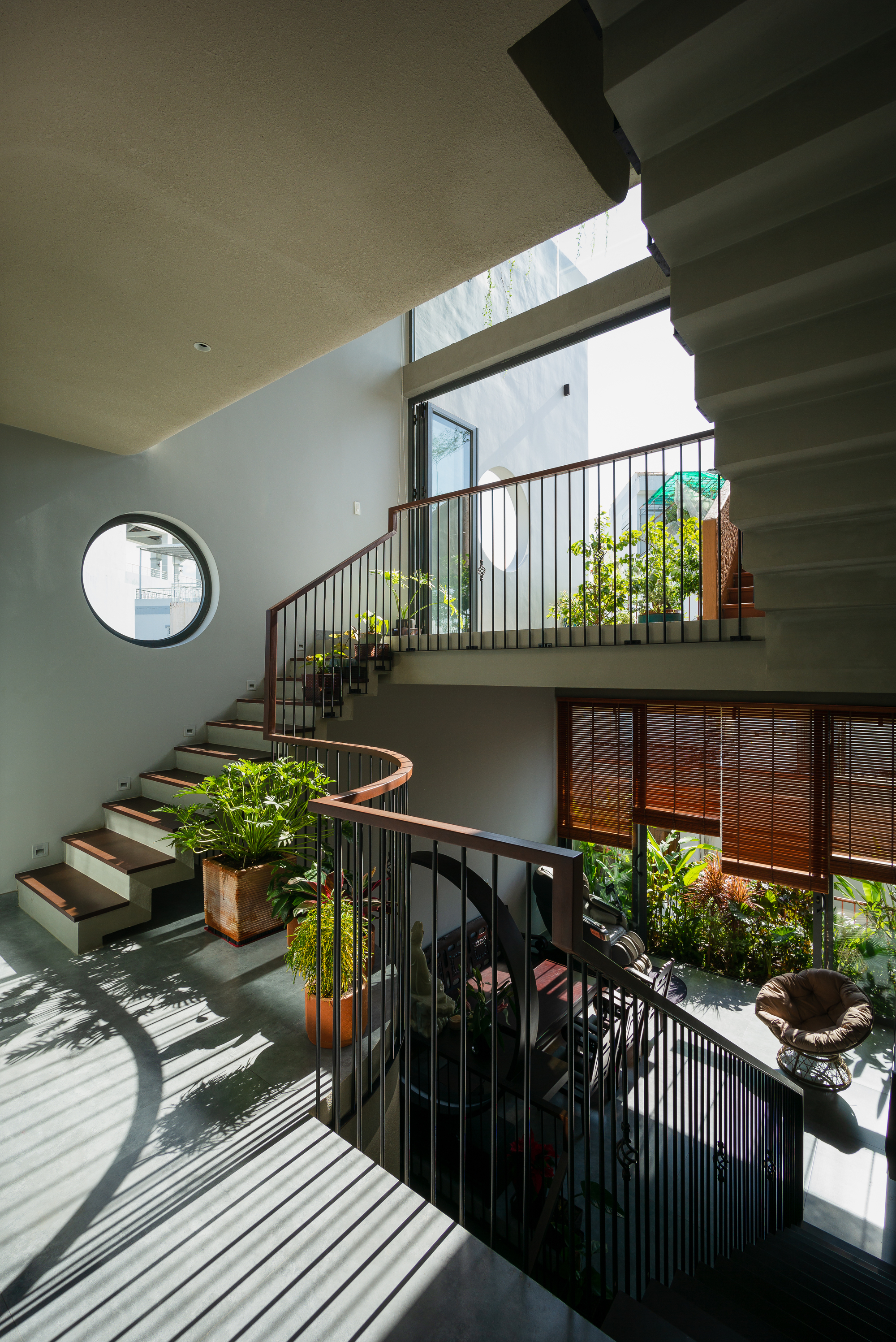 Triple-height stairwell with curved concrete steps, metal railings, and planted terraces receiving dappled sunlight from skylights