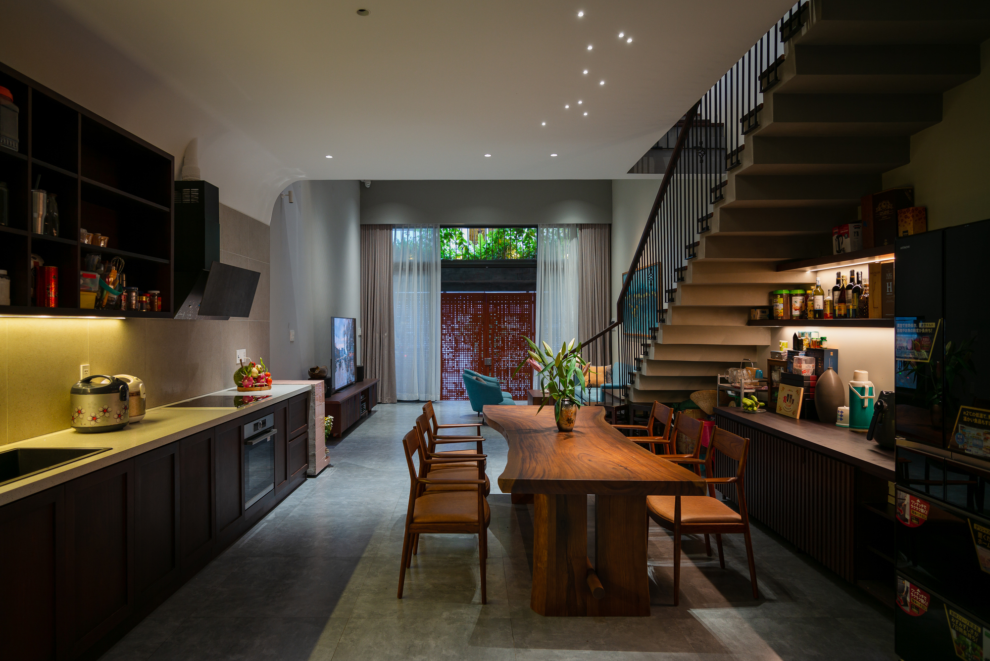 Open kitchen and dining space with timber table, floating staircase, and view to planted courtyard at dusk