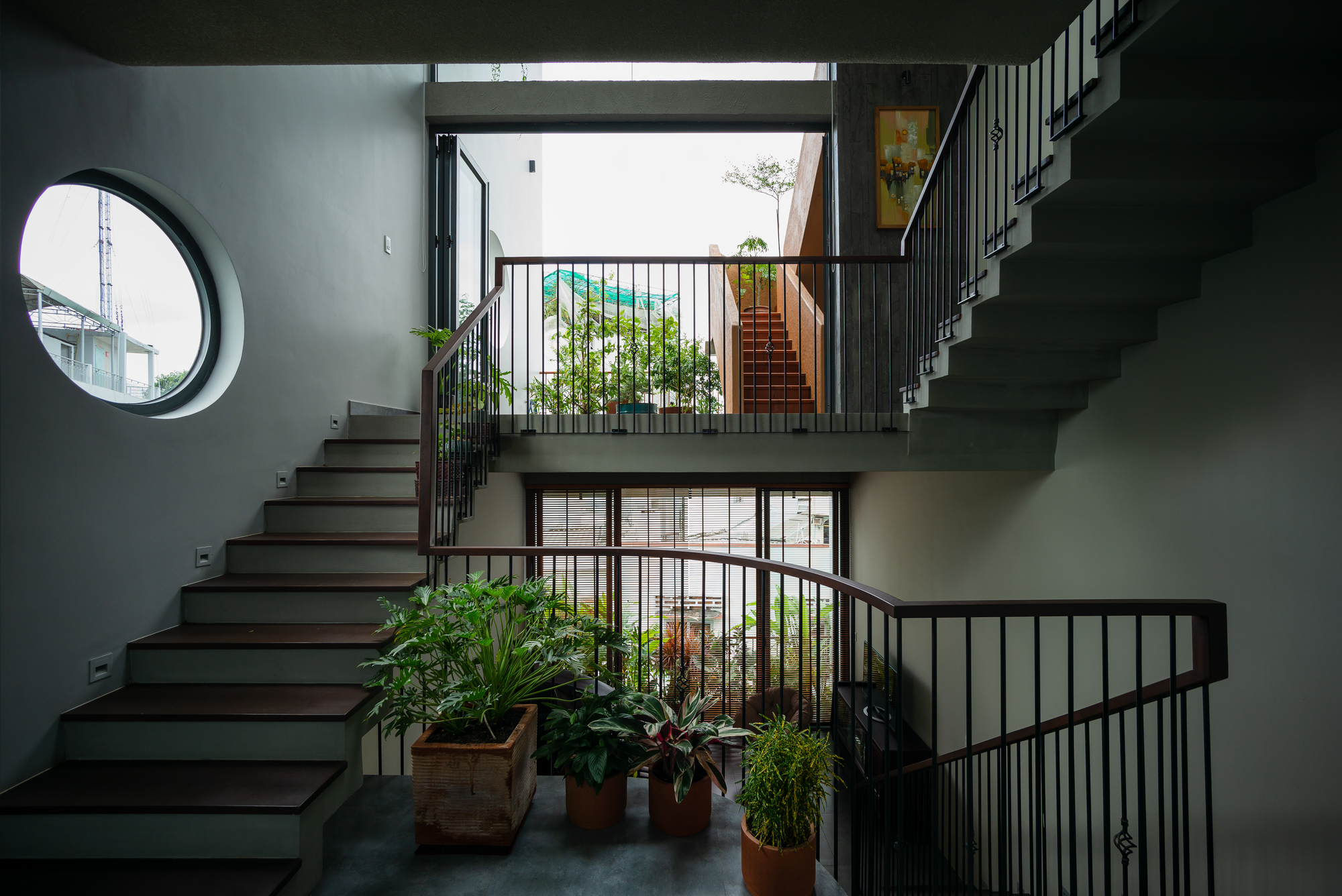 Multi-level staircase with circular window, metal balustrades, and potted plants on concrete landings in natural light