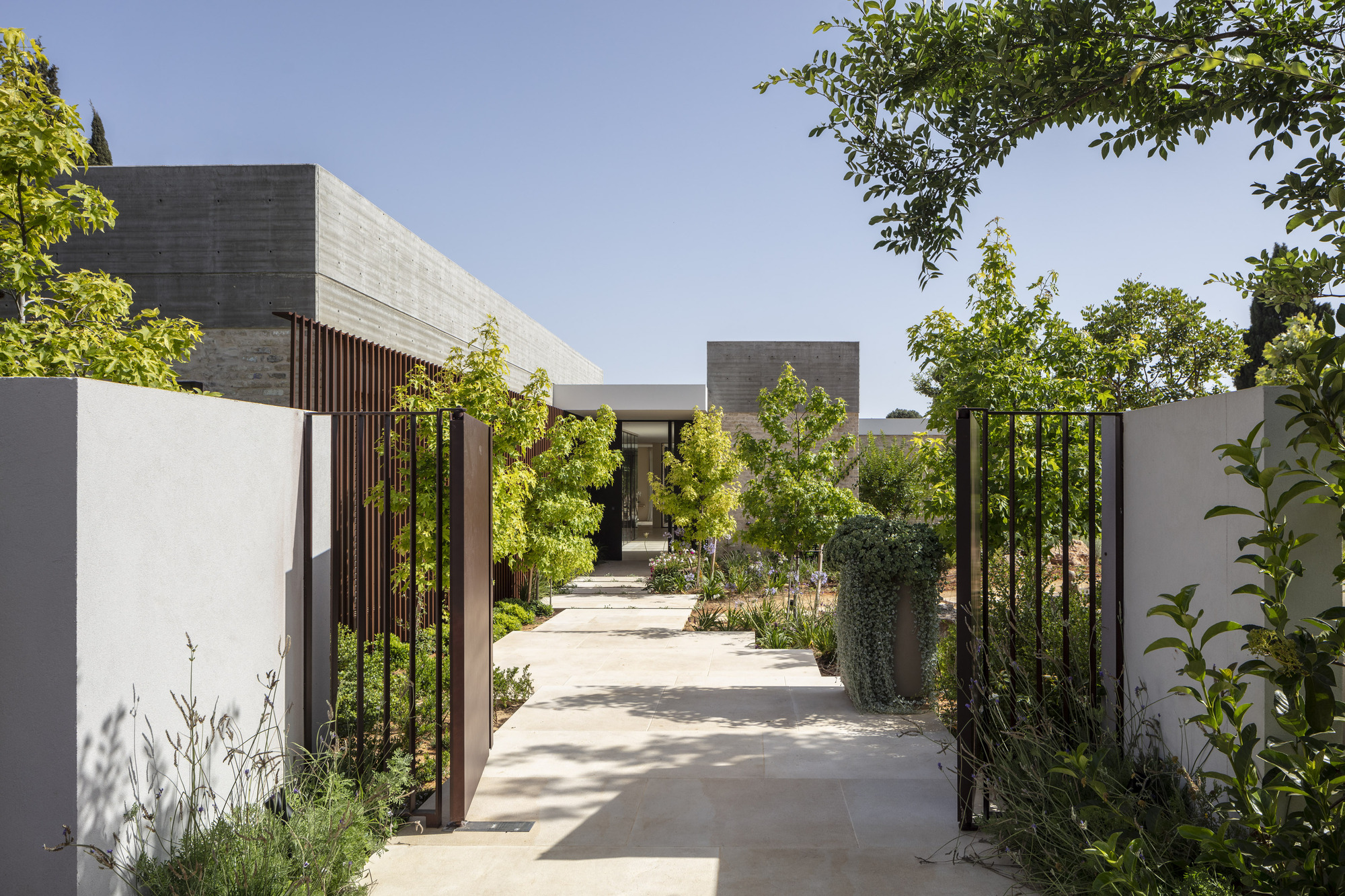 Entry pathway lined with white walls and vertical metal slats leading to concrete volumes under clear sky
