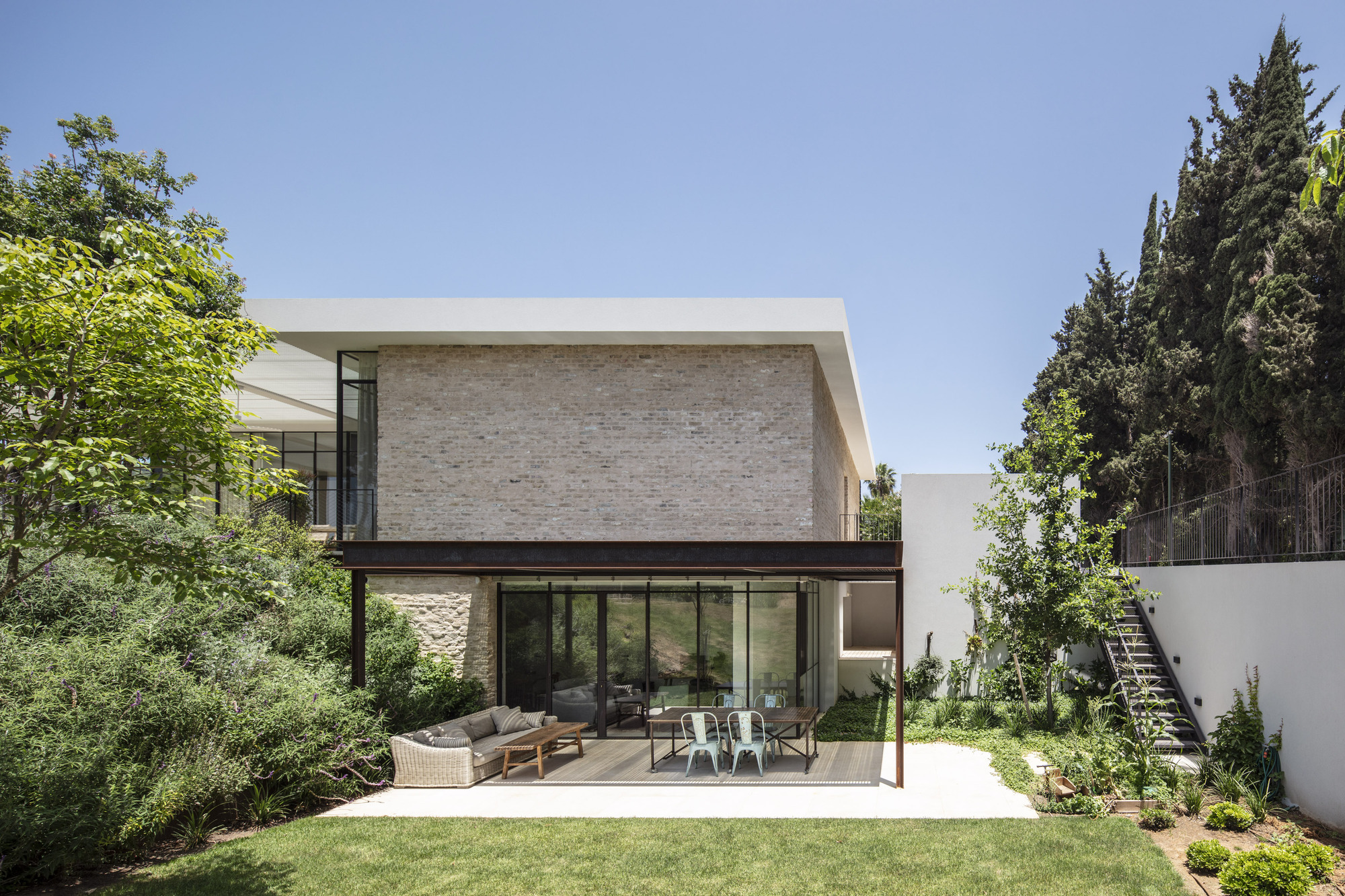 Garden view of the two-story volume with light brick cladding above a glazed terrace with outdoor furniture
