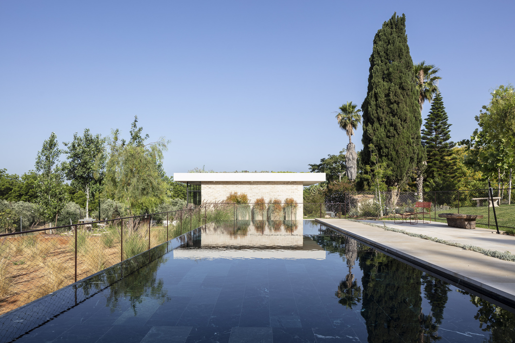 Stone pool pavilion with planted roof reflecting in the dark water under a clear summer sky