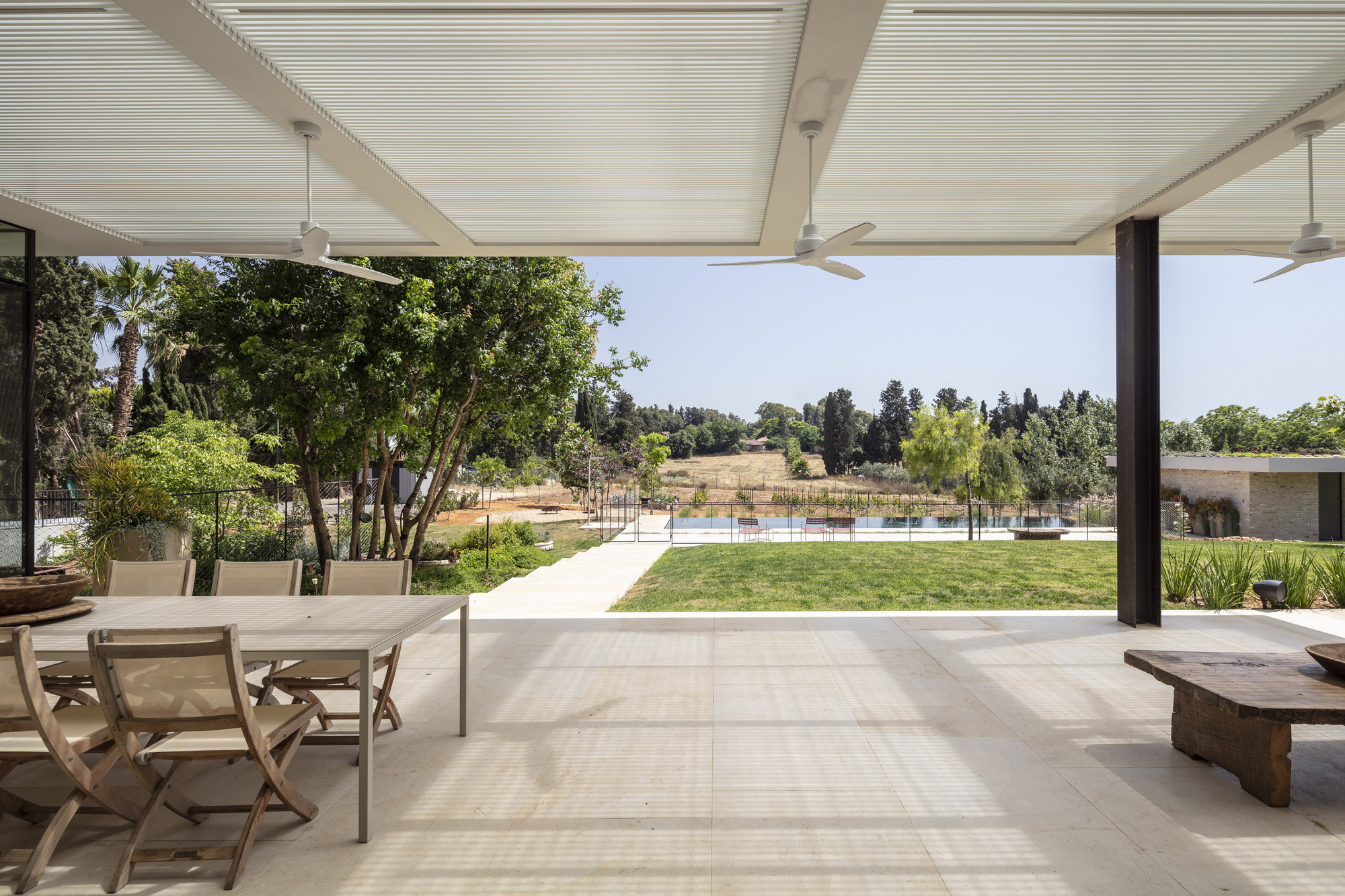 Covered terrace with slatted ceiling and ceiling fans opening onto lawn and pool beyond mature trees