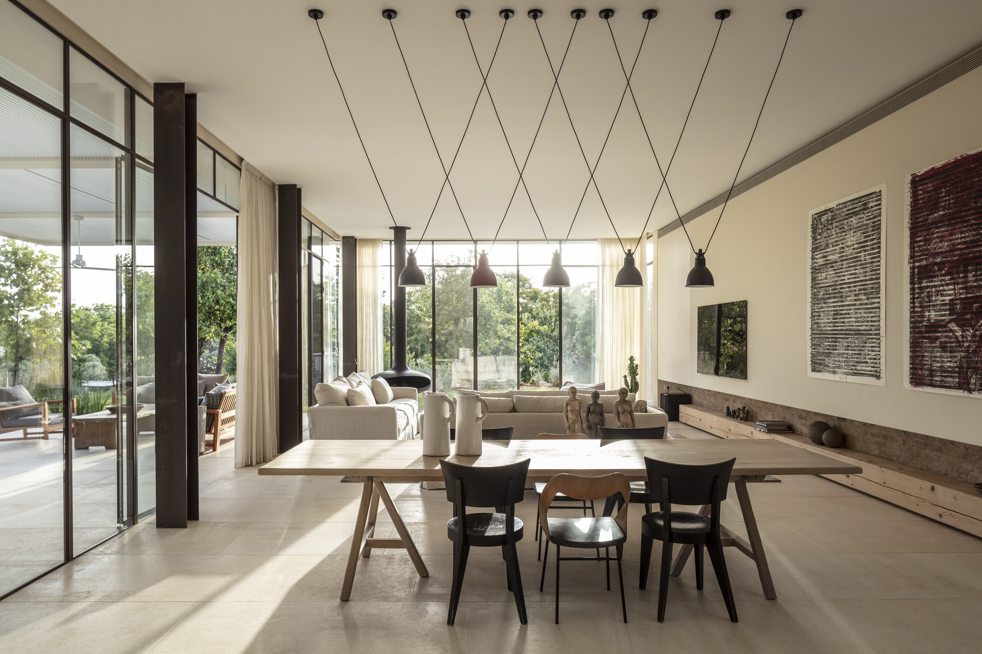 Open living and dining area with pendant lights on crossed cables and floor-to-ceiling glazing overlooking trees