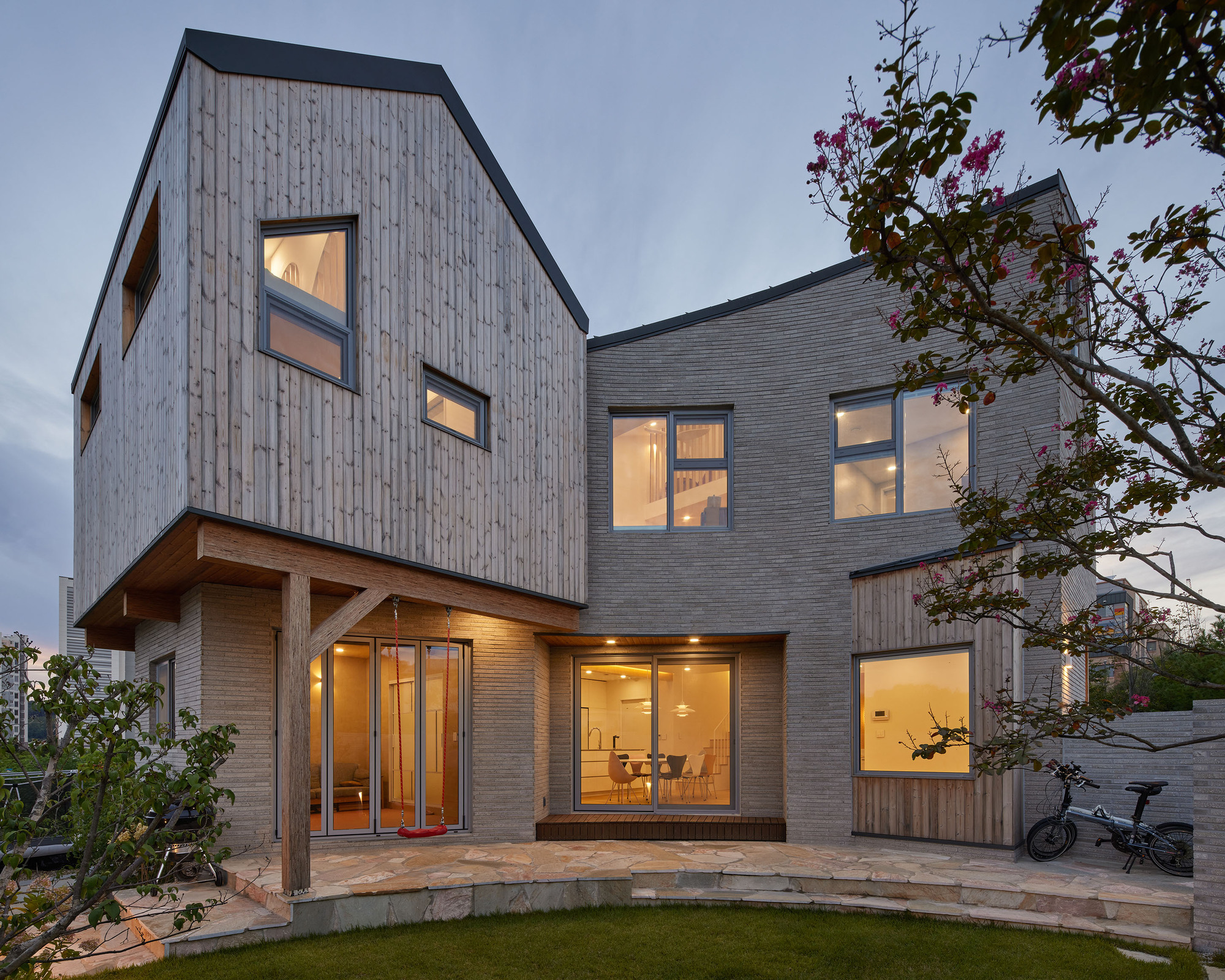 Rear courtyard showing glazed openings with warm interior light at dusk framed by flowering trees